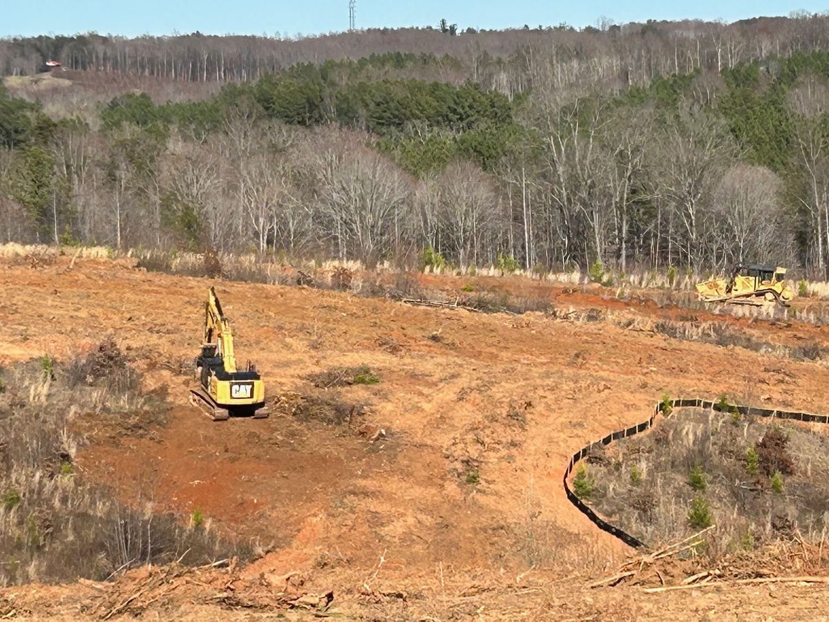 Excavator on a cleared, red-soiled construction site, trees in background under a blue sky.