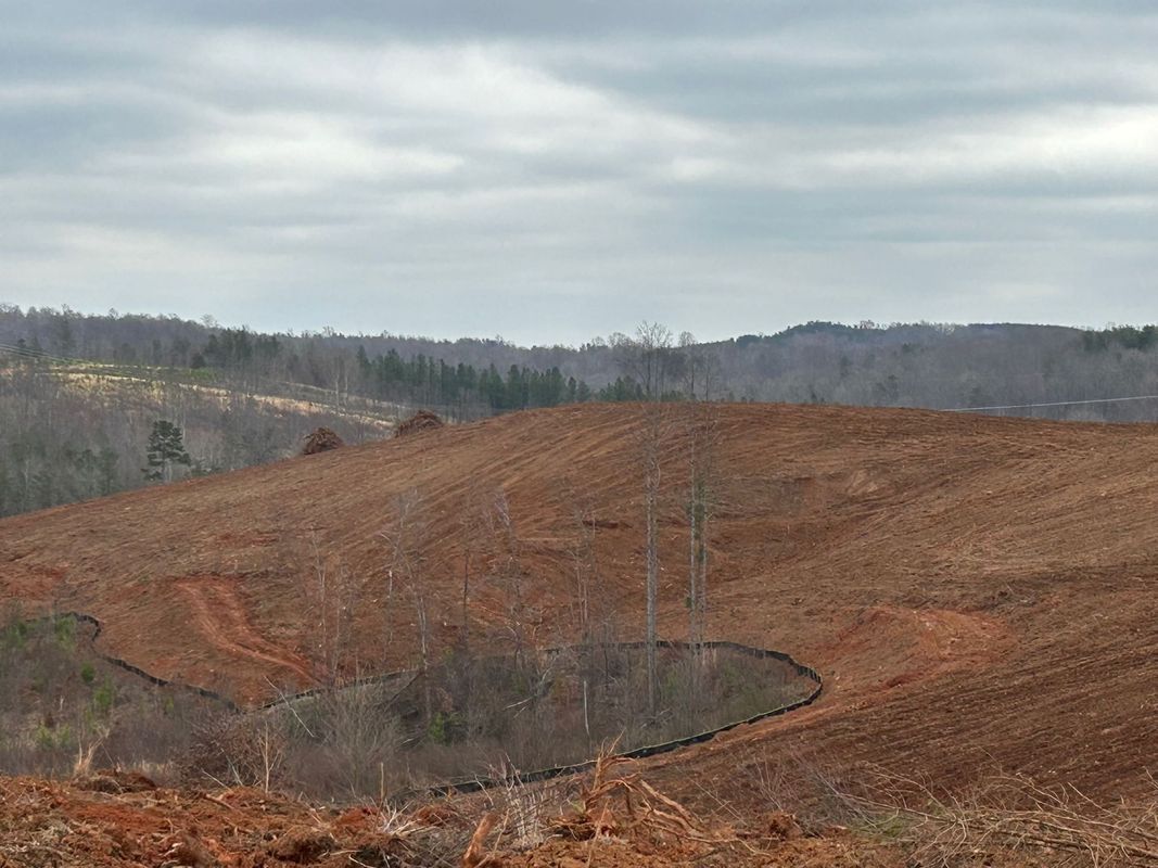 Cleared red dirt land with power lines, hills, and a cloudy sky.