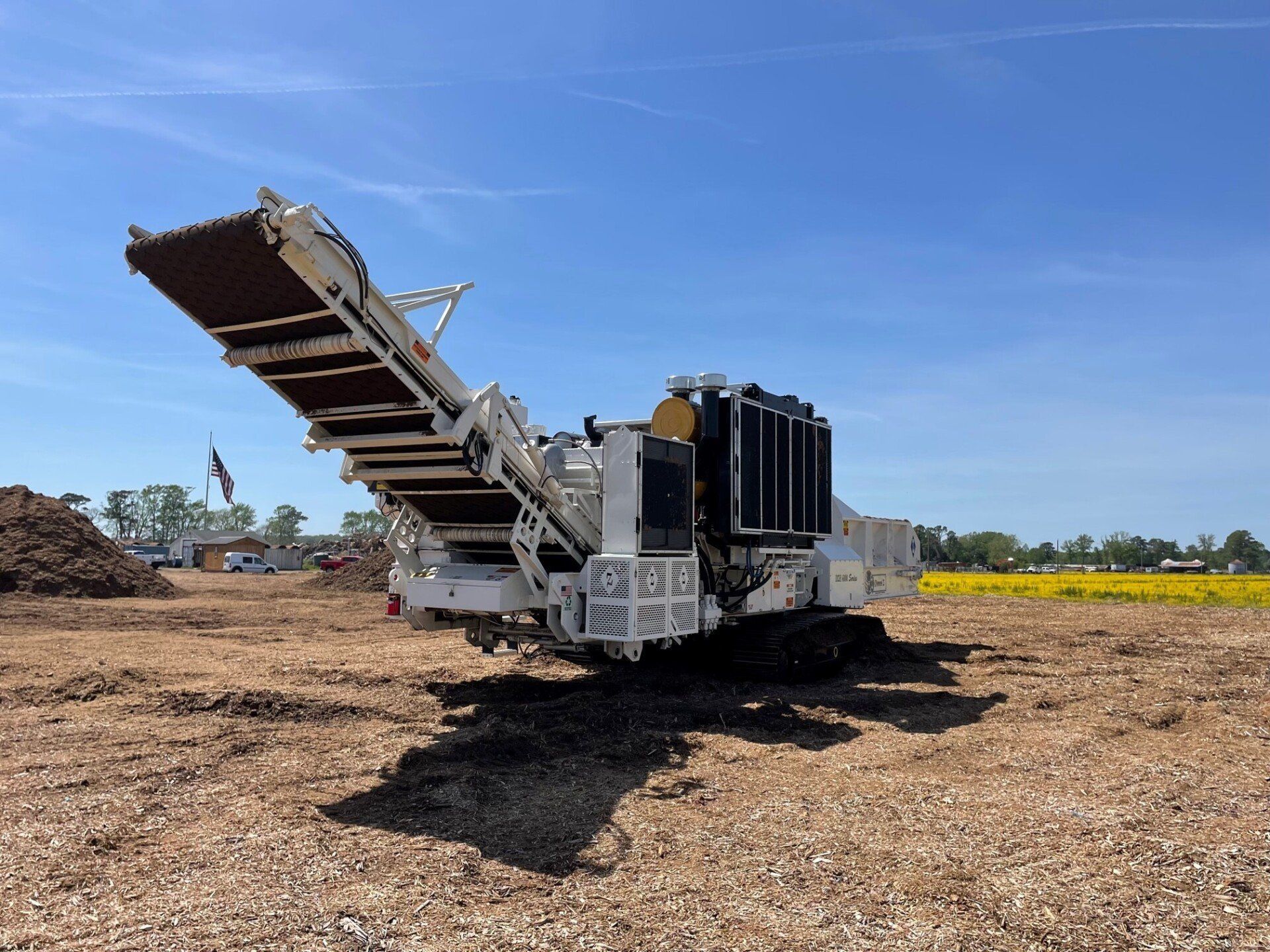 White industrial machine with conveyor belt in a field, under a blue sky.