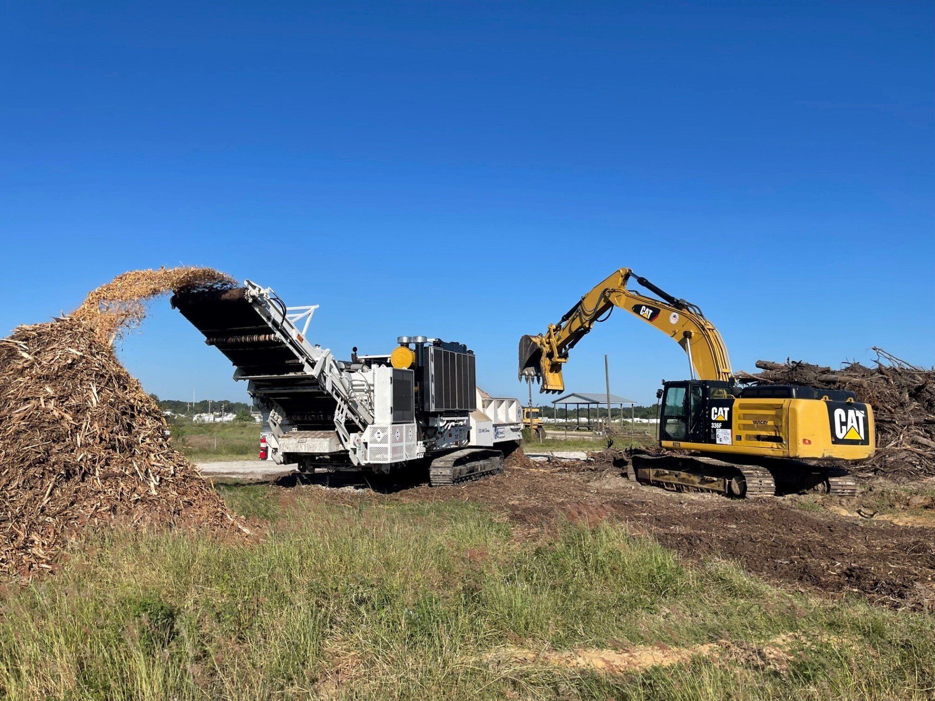 Excavator feeding wood debris into a wood grinder; mulch pile on the left, blue sky.