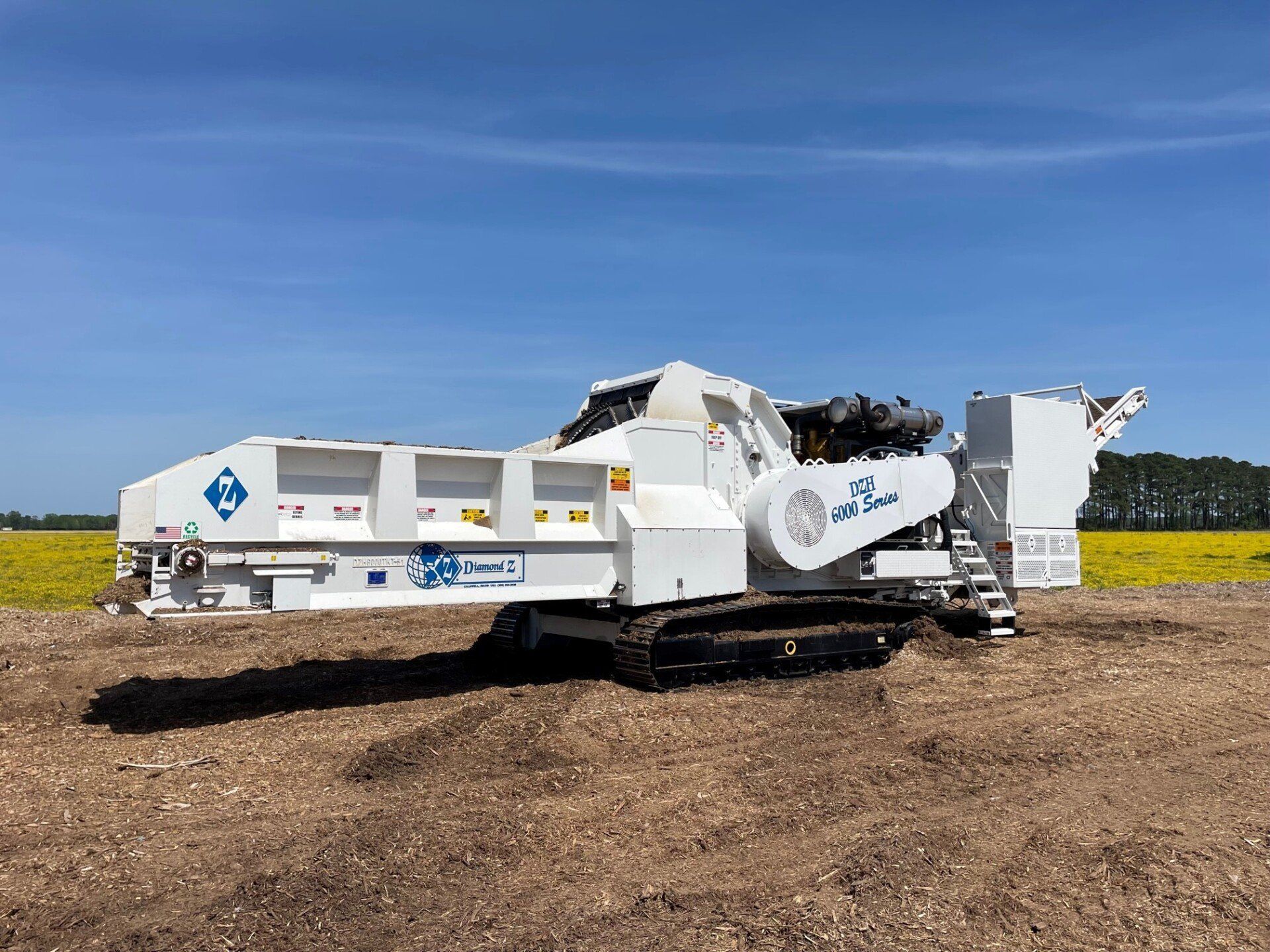 White industrial crushing machine on a dirt field with a yellow flower field in the background, blue sky.