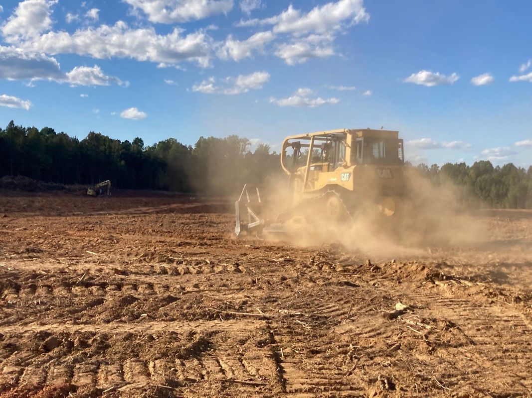 A yellow bulldozer kicking up dust while leveling a brown field with trees in the background under a blue sky.