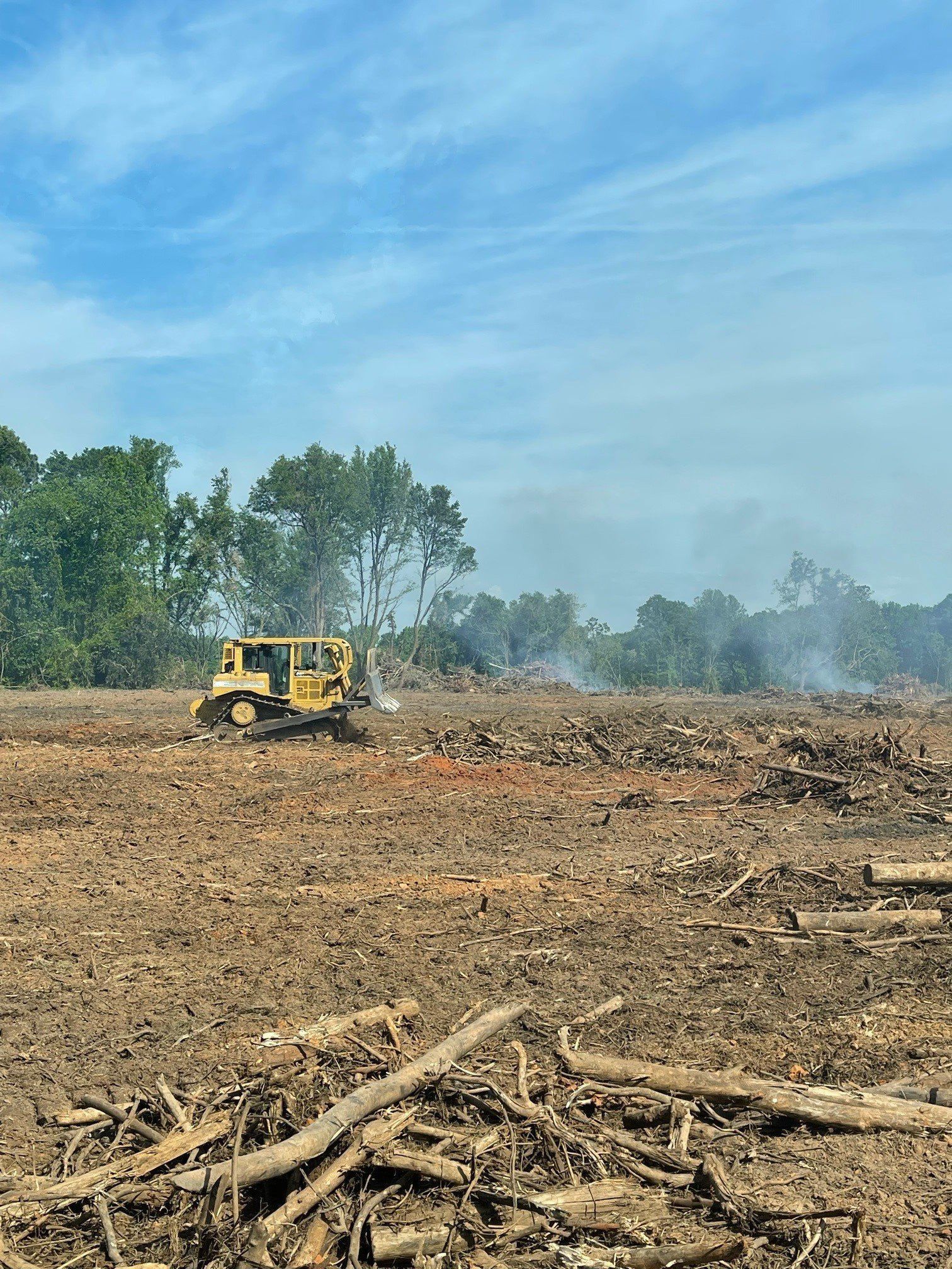 Yellow bulldozer clearing a muddy field, with cut trees and smoke in the background. Blue sky with clouds.