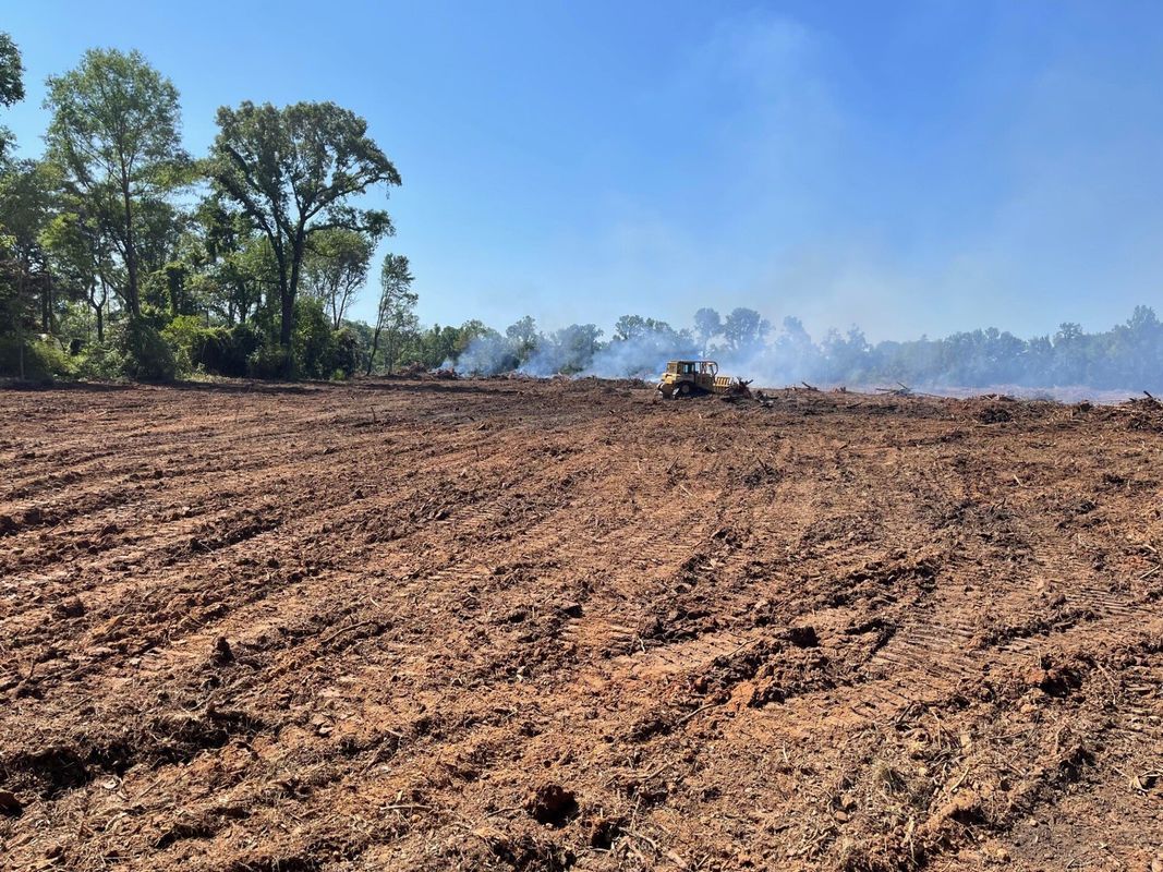 Brown, tilled field with a bulldozer amid smoke; trees line the background under a blue sky.