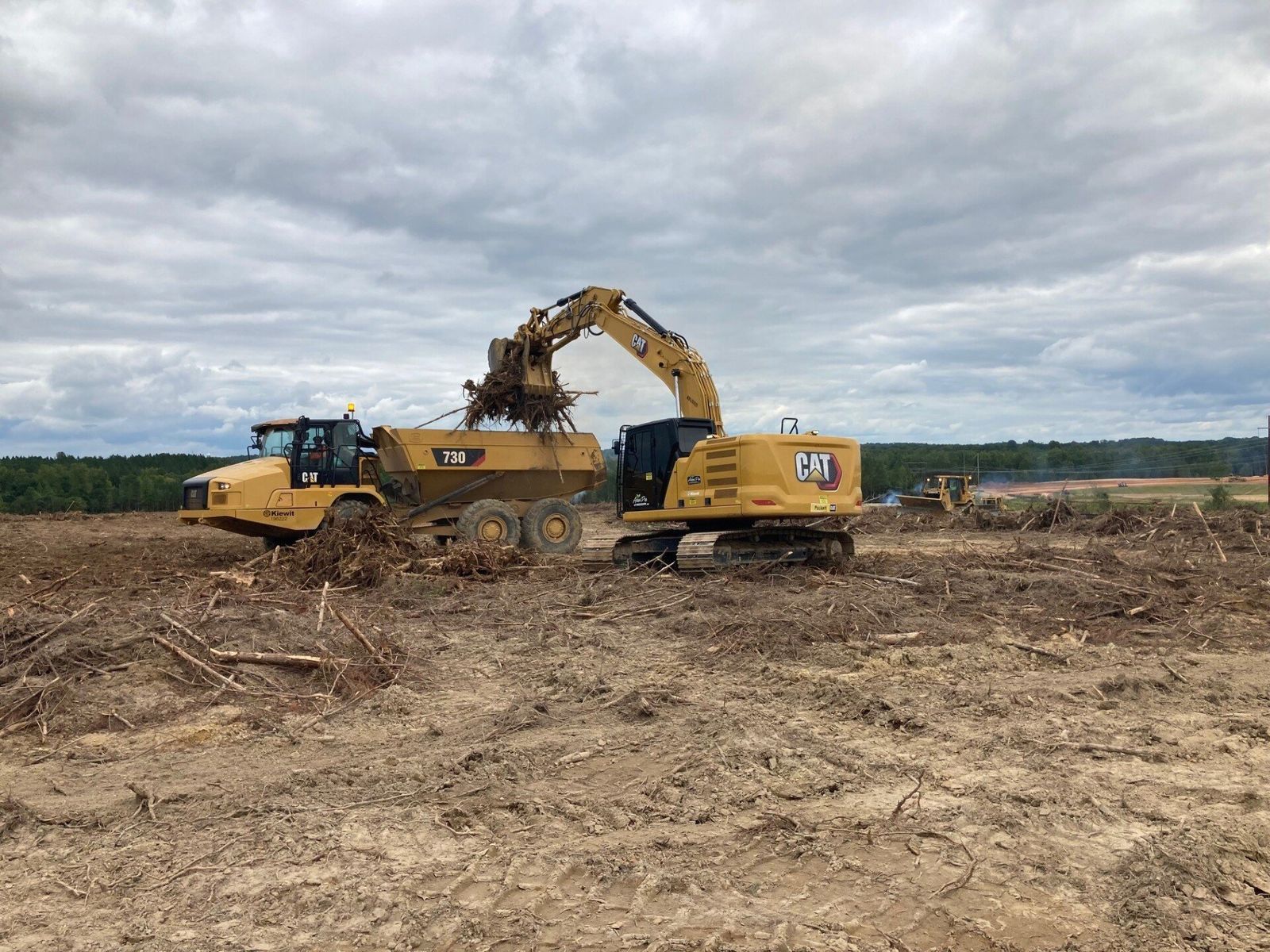 Yellow excavator loading debris into a dump truck in a cleared field under a cloudy sky.