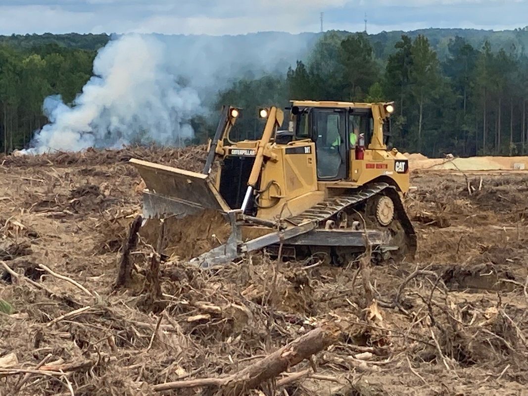 Yellow bulldozer clearing a field of debris, with smoke in the background; a forest visible in the distance.