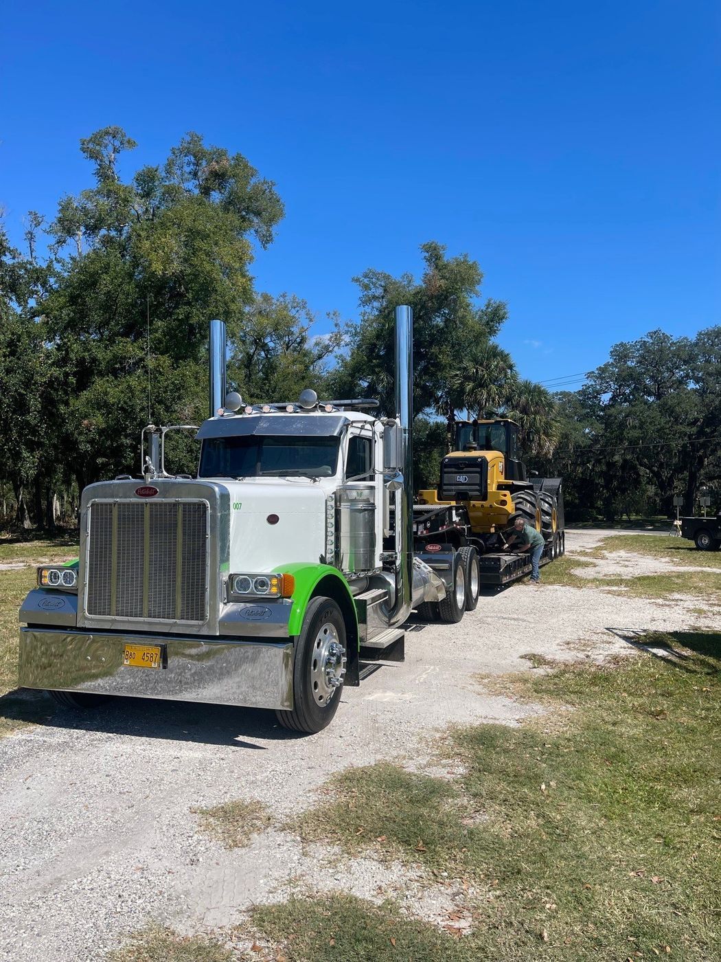 White and green semi-truck hauling a yellow tractor on a trailer, parked on a gravel drive.