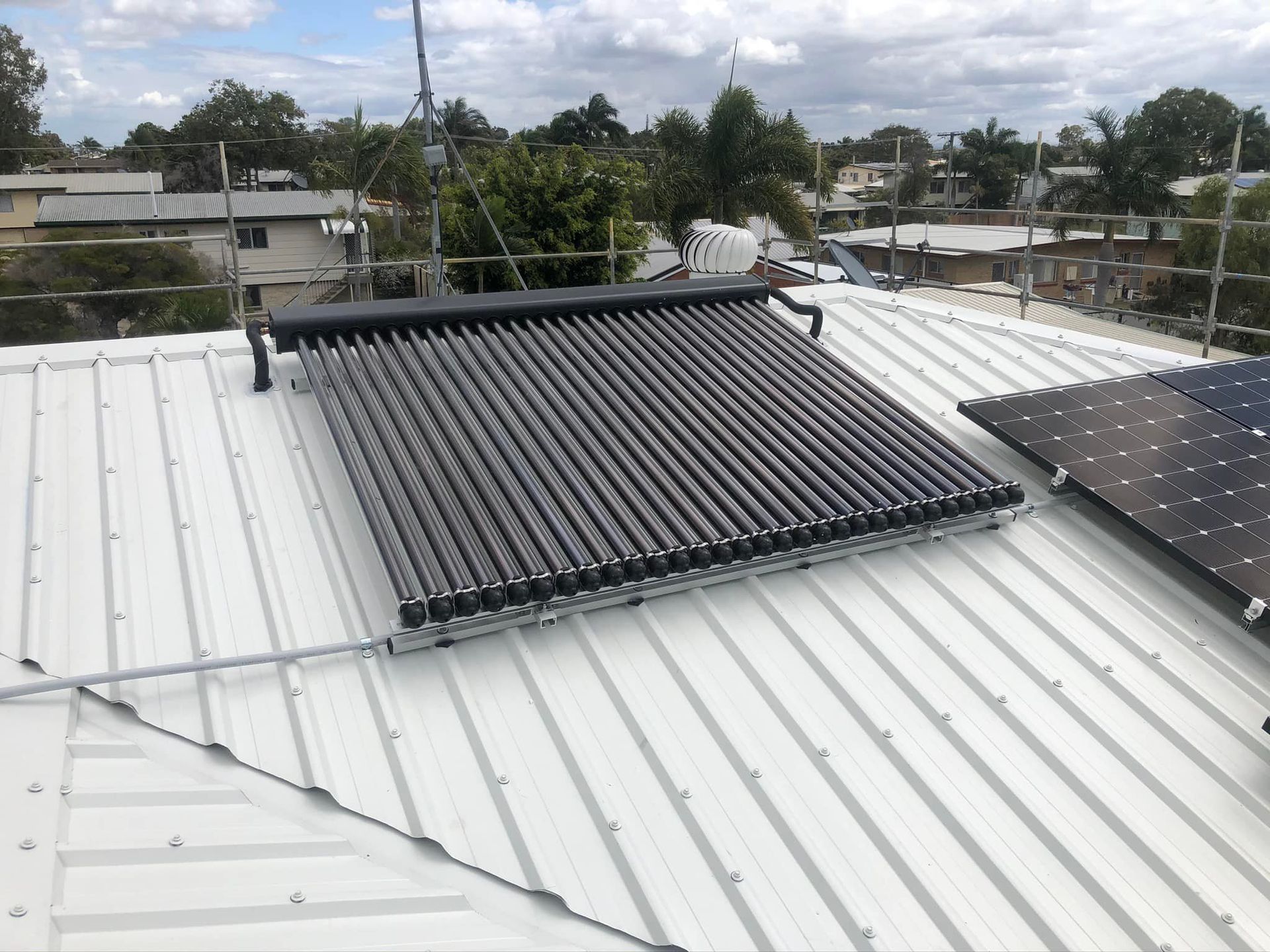 A solar water heater with rows of dark glass tubes sits on a white metal roof next to a solar panel — Hales Plumbing & Roofing In Gracemere, QLD