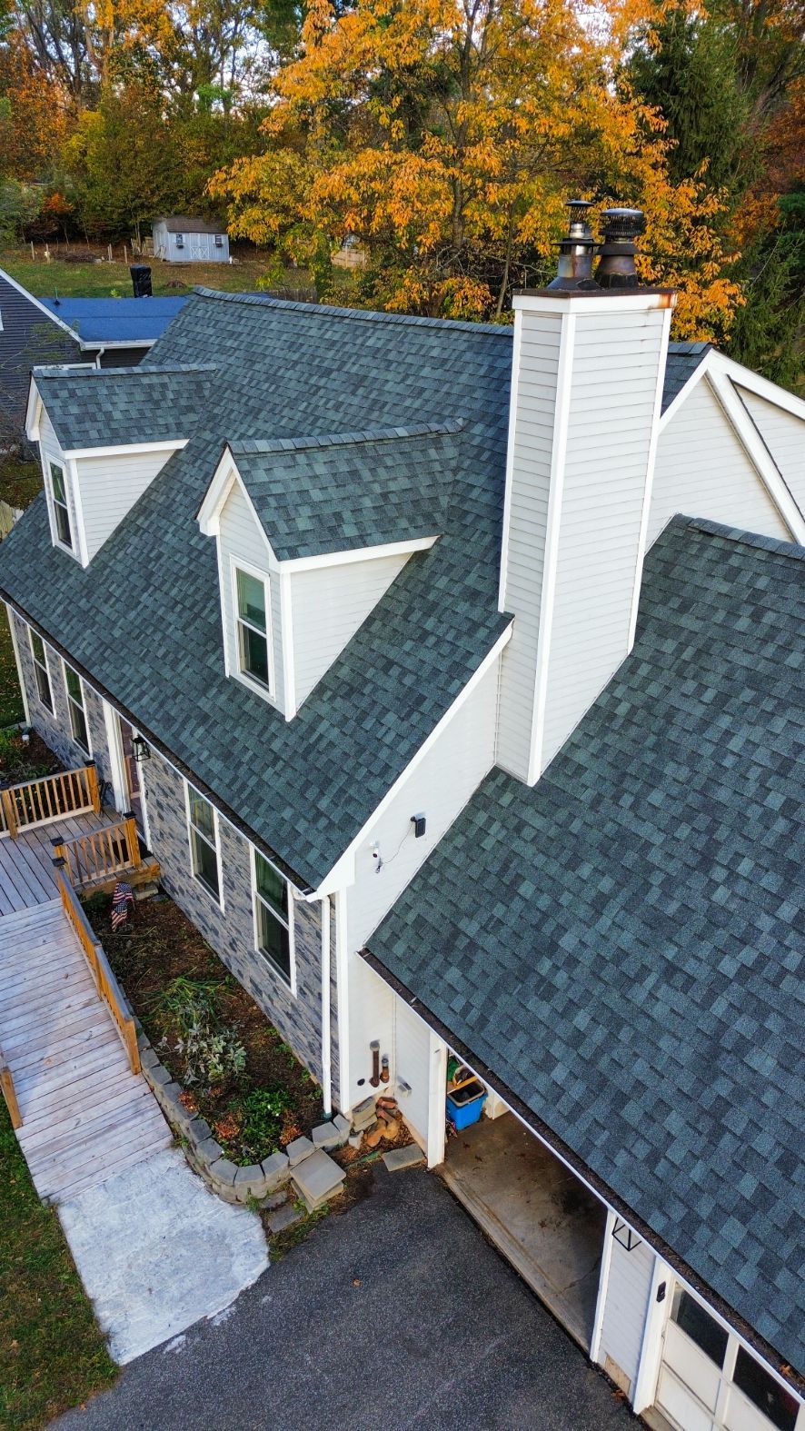 Aerial view of a gray-shingled house with white trim and a chimney, surrounded by autumn trees.