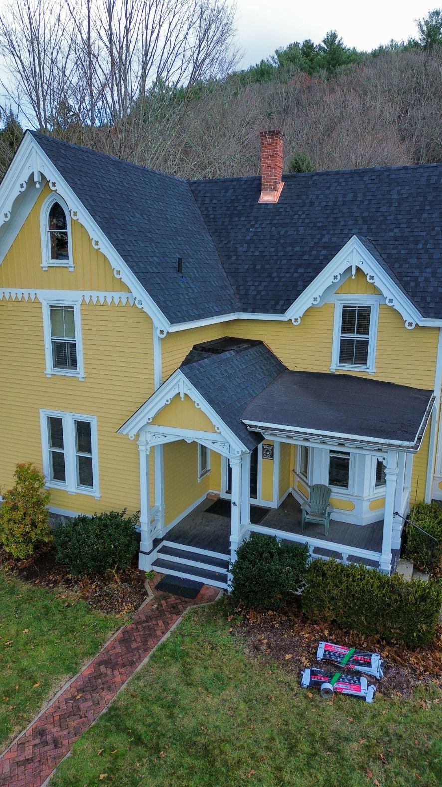 Yellow Victorian house with dark roof, white trim, brick chimney, and front porch in a wooded yard