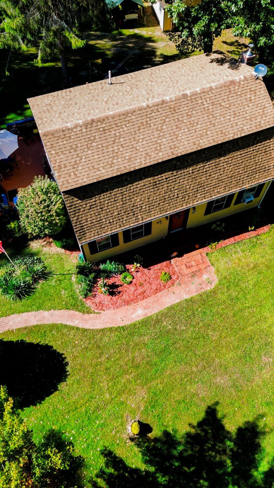 Aerial view of a small tan-roofed house with a red brick patio, surrounded by green lawn and trees.