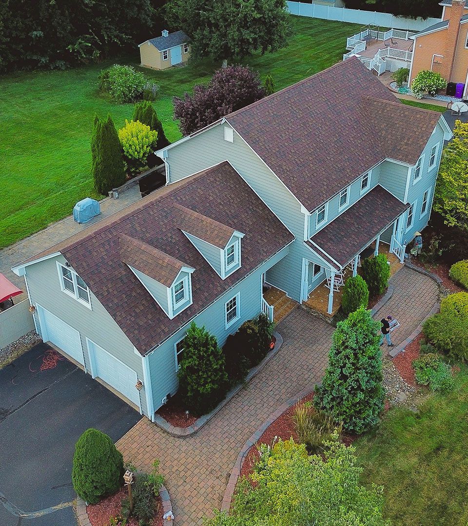 Aerial view of a two-story blue house with a brown roof, dormers, and a paved driveway surrounded by green landscaping.