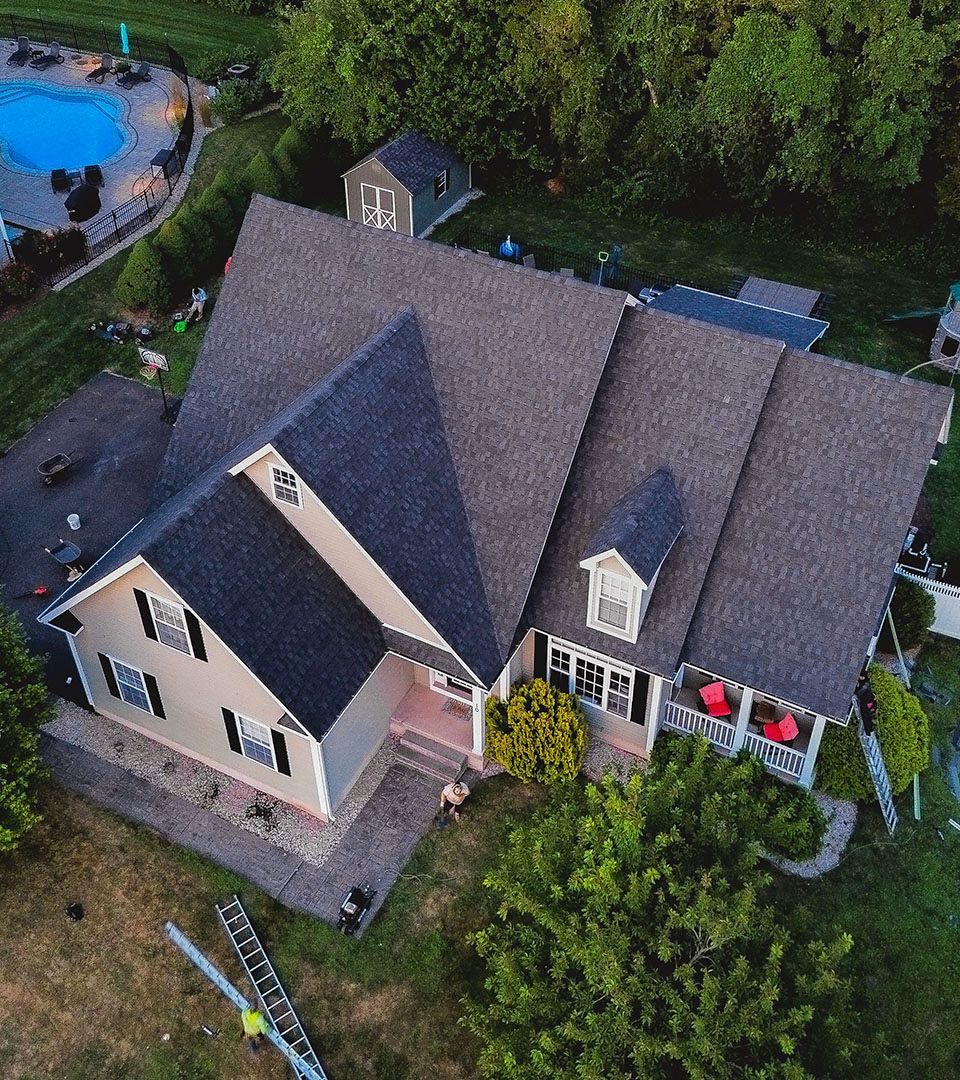Aerial view of a beige house with a dark gray roof, pool, and lush green surroundings.