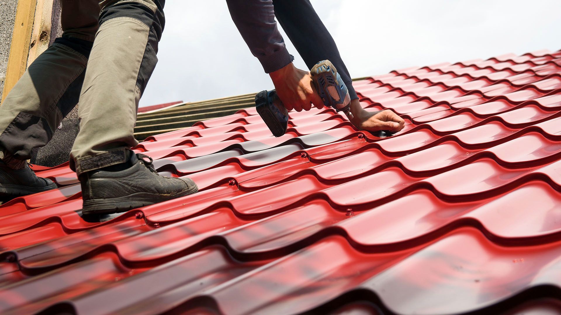 Construction workers installing bright red metal roof panels using a drill.