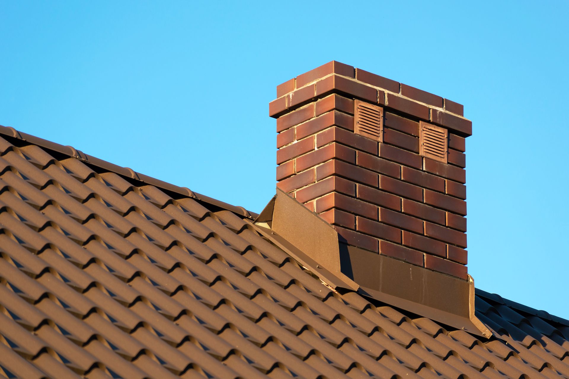 Brown brick chimney on a brown tile roof against a clear blue sky.