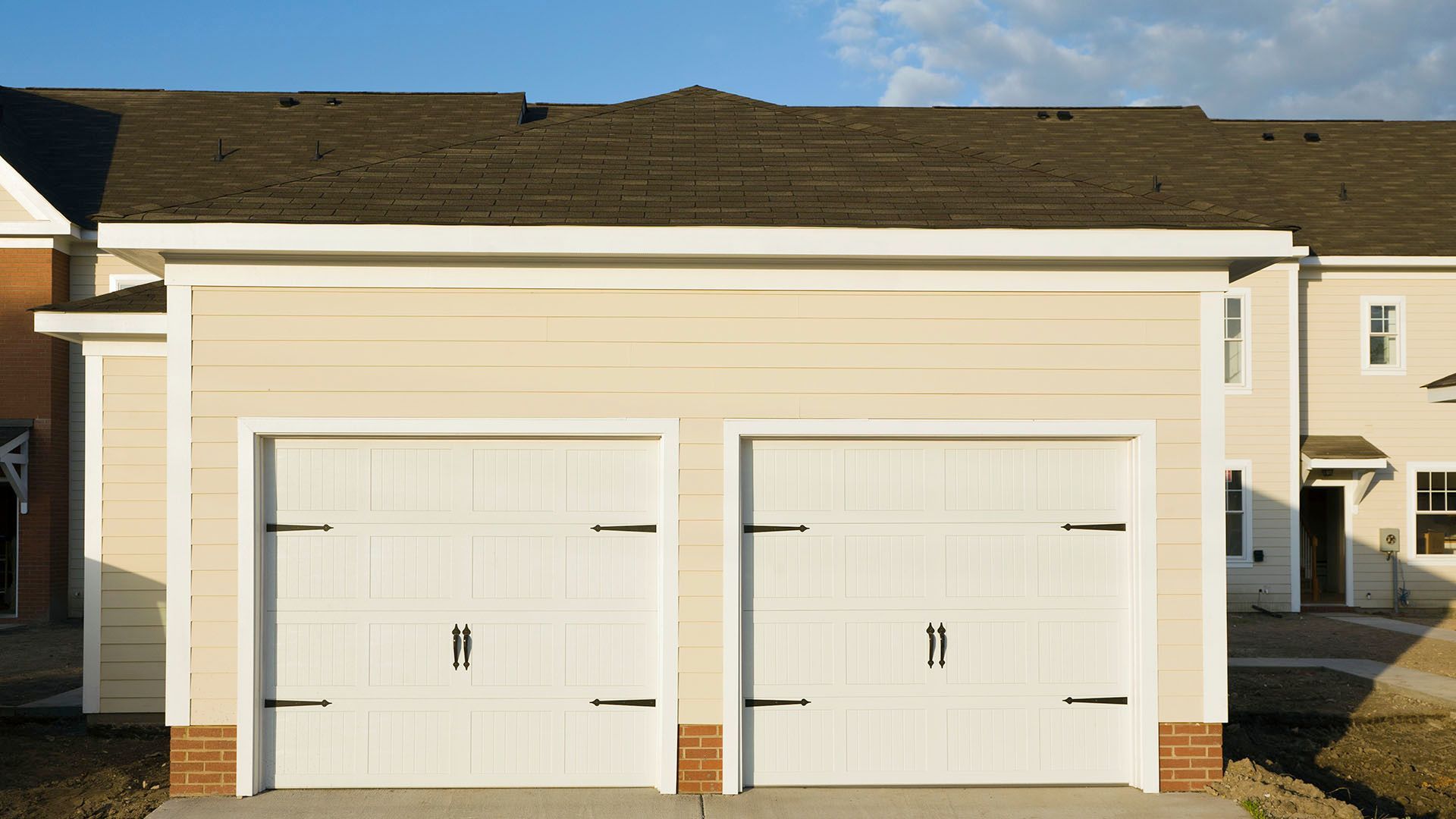 Two white garage doors with black hardware, tan siding, brick base.