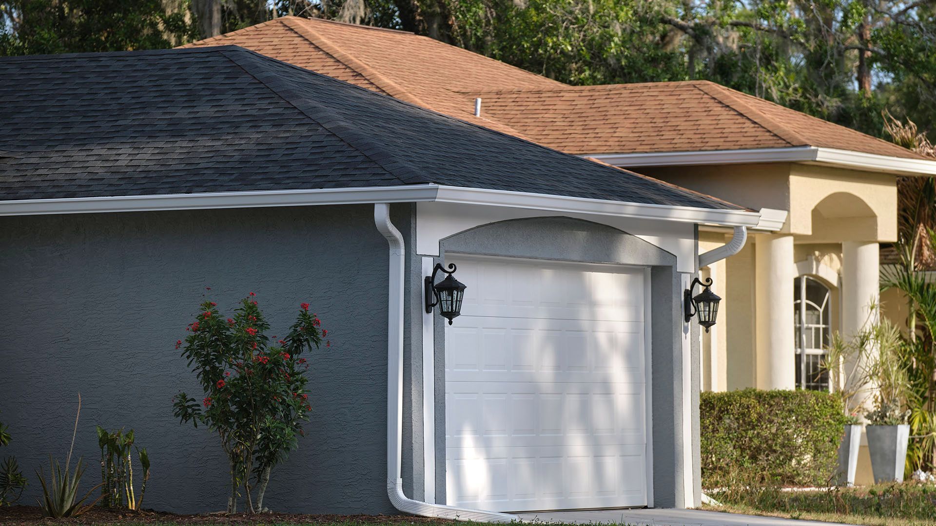 Garage and house exterior, blue-gray garage with white door, black lamps, brown and beige roof, green bushes, sunny day.