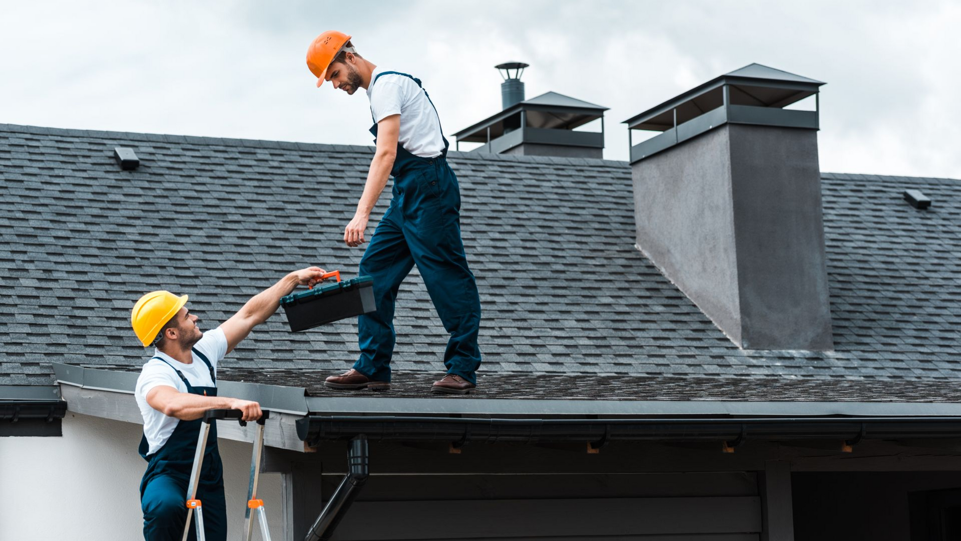 Two roofers in hard hats on a rooftop. One hands a toolbox to the other.