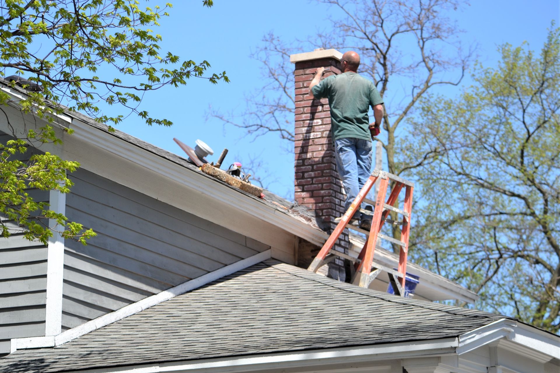 Person on ladder repairing a brick chimney on a house roof. Blue sky.