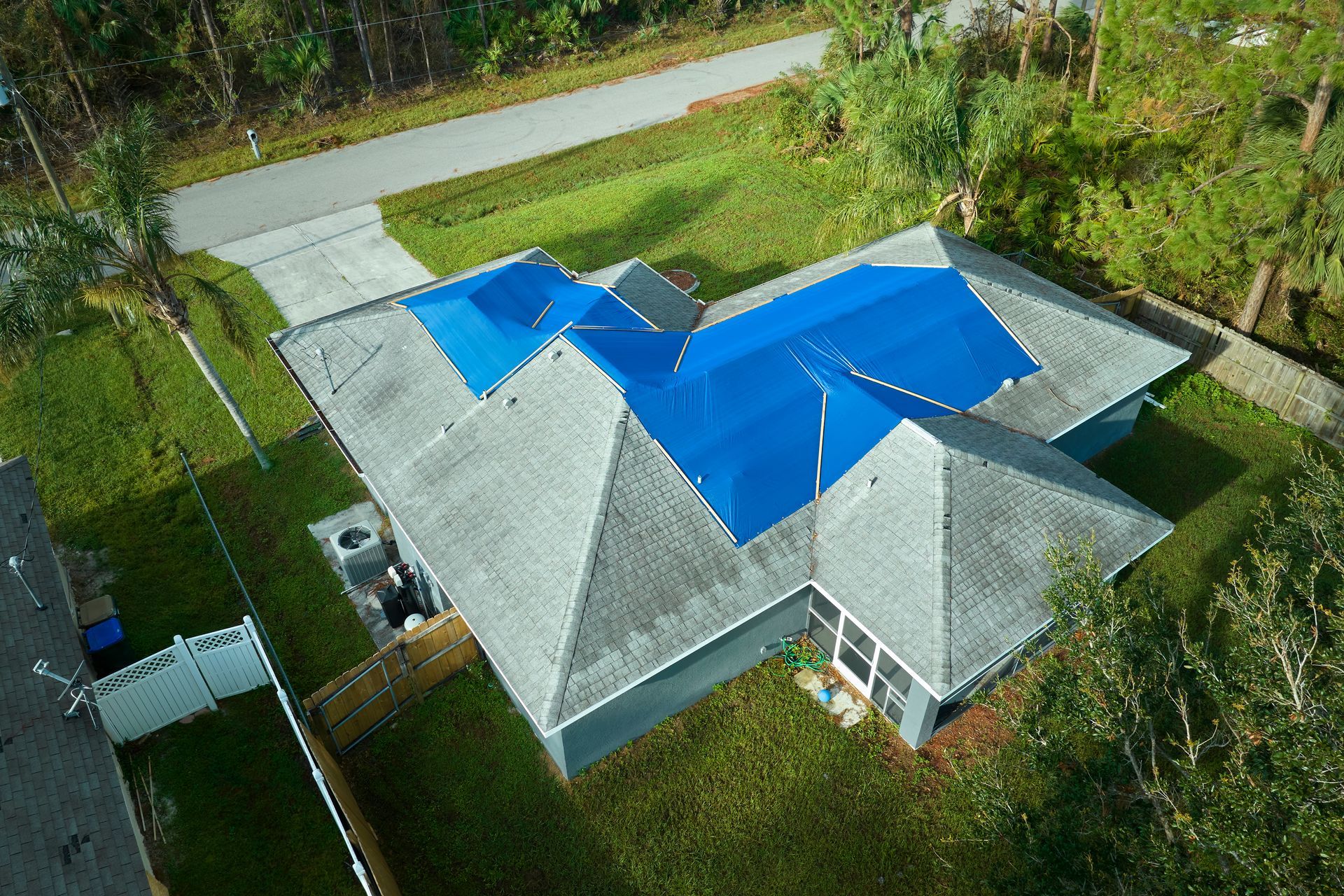 Aerial view of a house with a gray roof and blue tarp covering sections. Driveway and green lawn surround the house.
