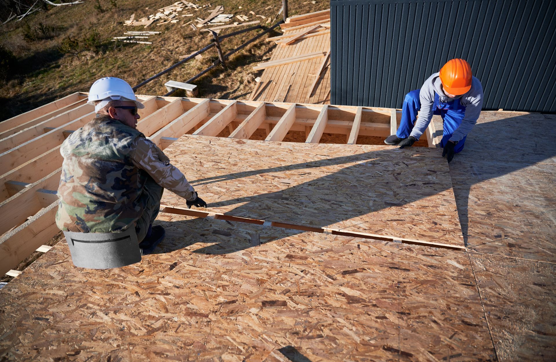 Two workers installing plywood roofing on a sloped roof, one in a white hard hat and one in orange.