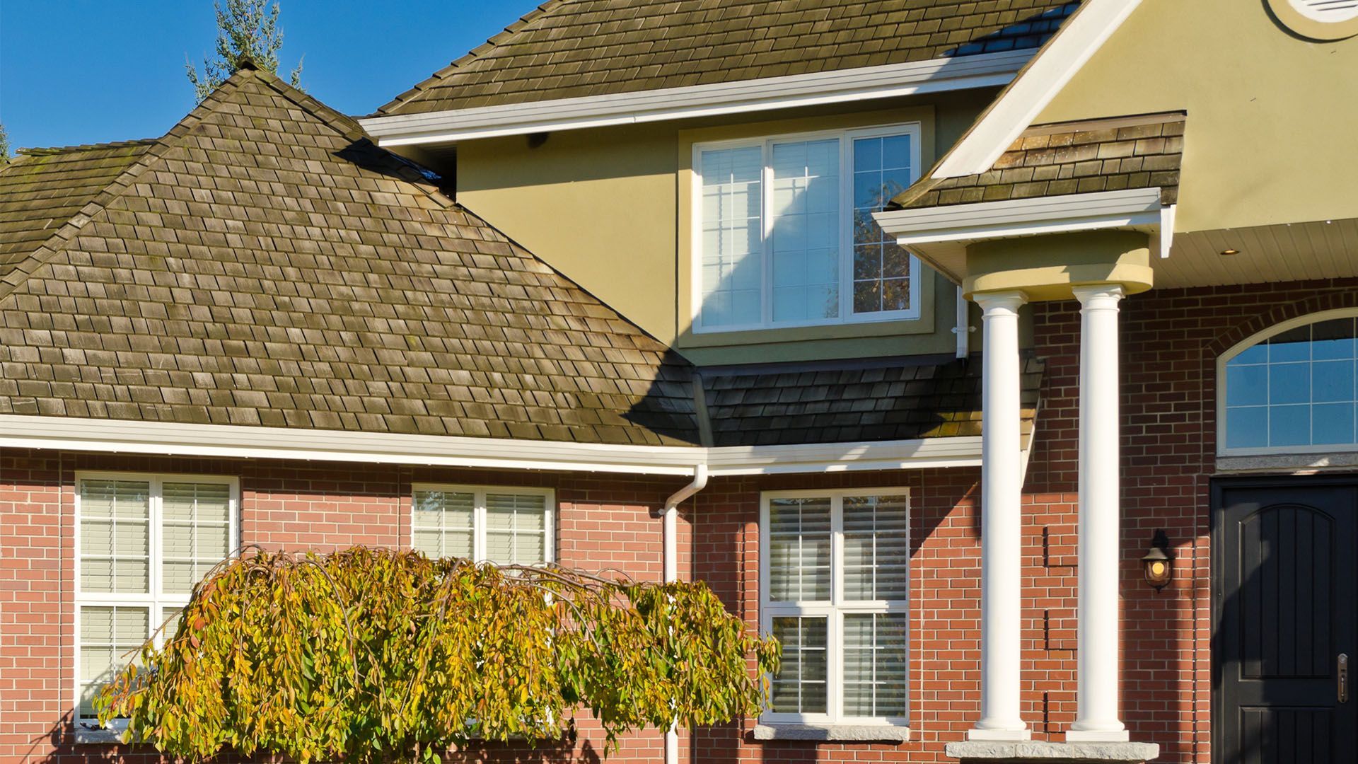 Brick house with multiple windows, columns, and a brown shingled roof. A small tree is in the foreground.