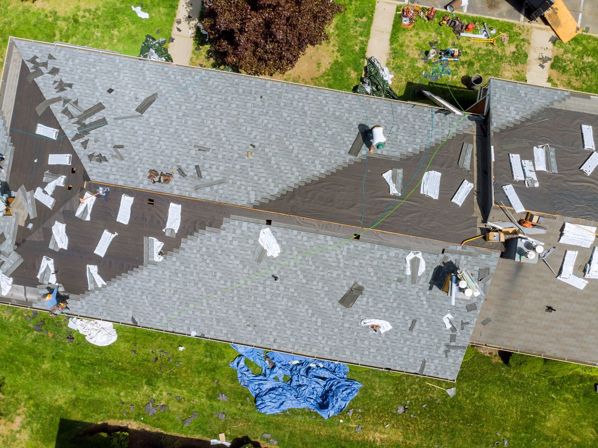 An aerial view of a house roof undergoing repairs, with sections of grey shingles partially removed and debris on the lawn.