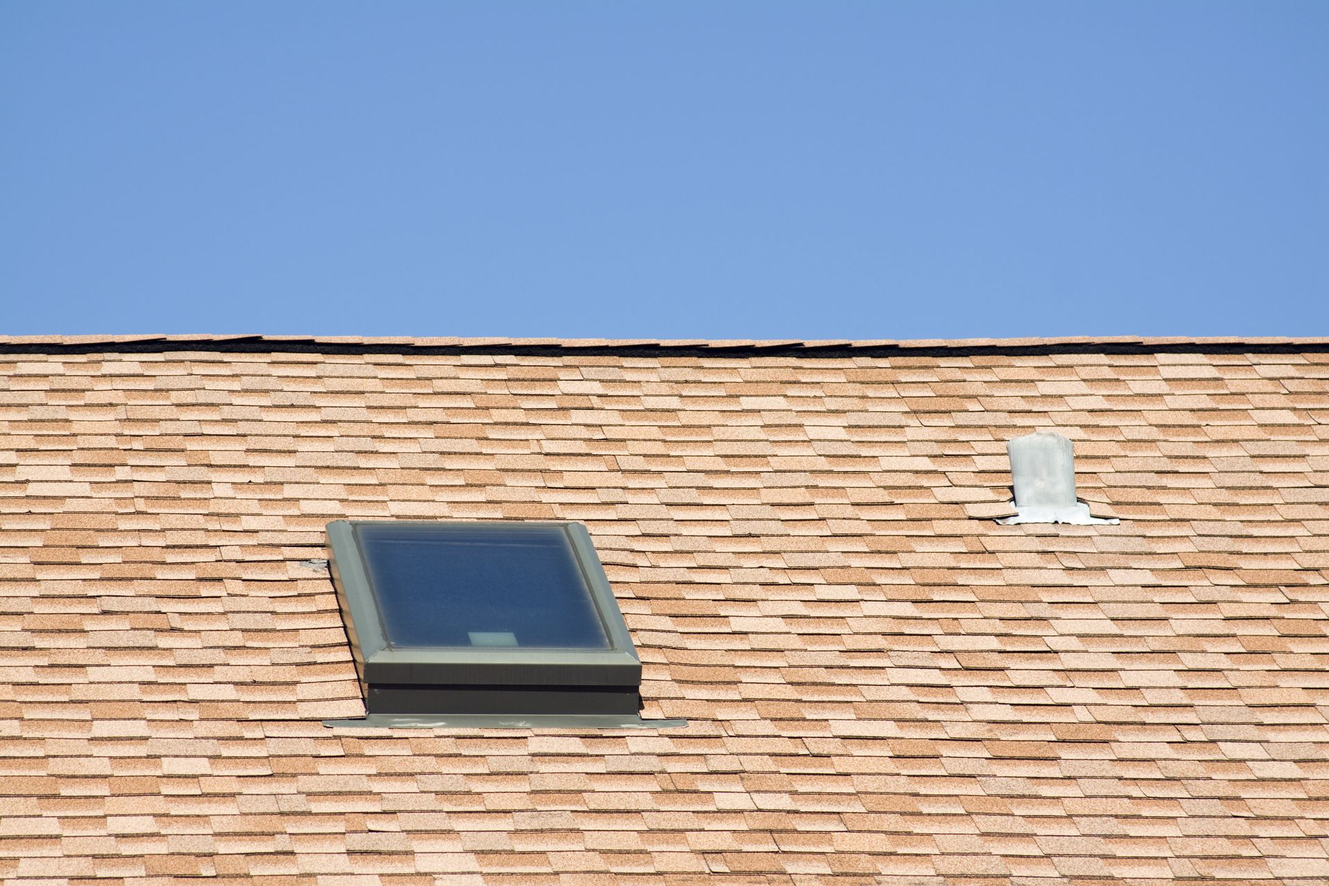 Brown shingled roof with a rectangular skylight and a small vent against a blue sky.