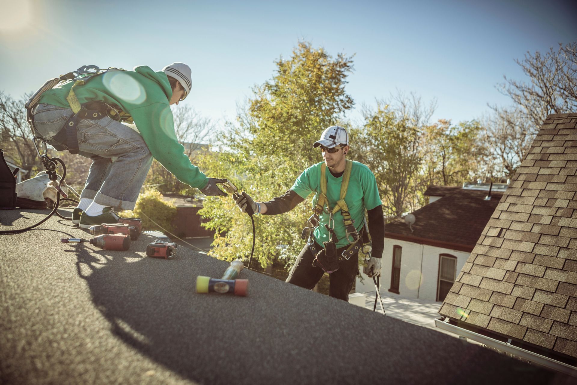 Two roofers in harnesses on a roof, handing objects. Sunny day, greenery in the background.