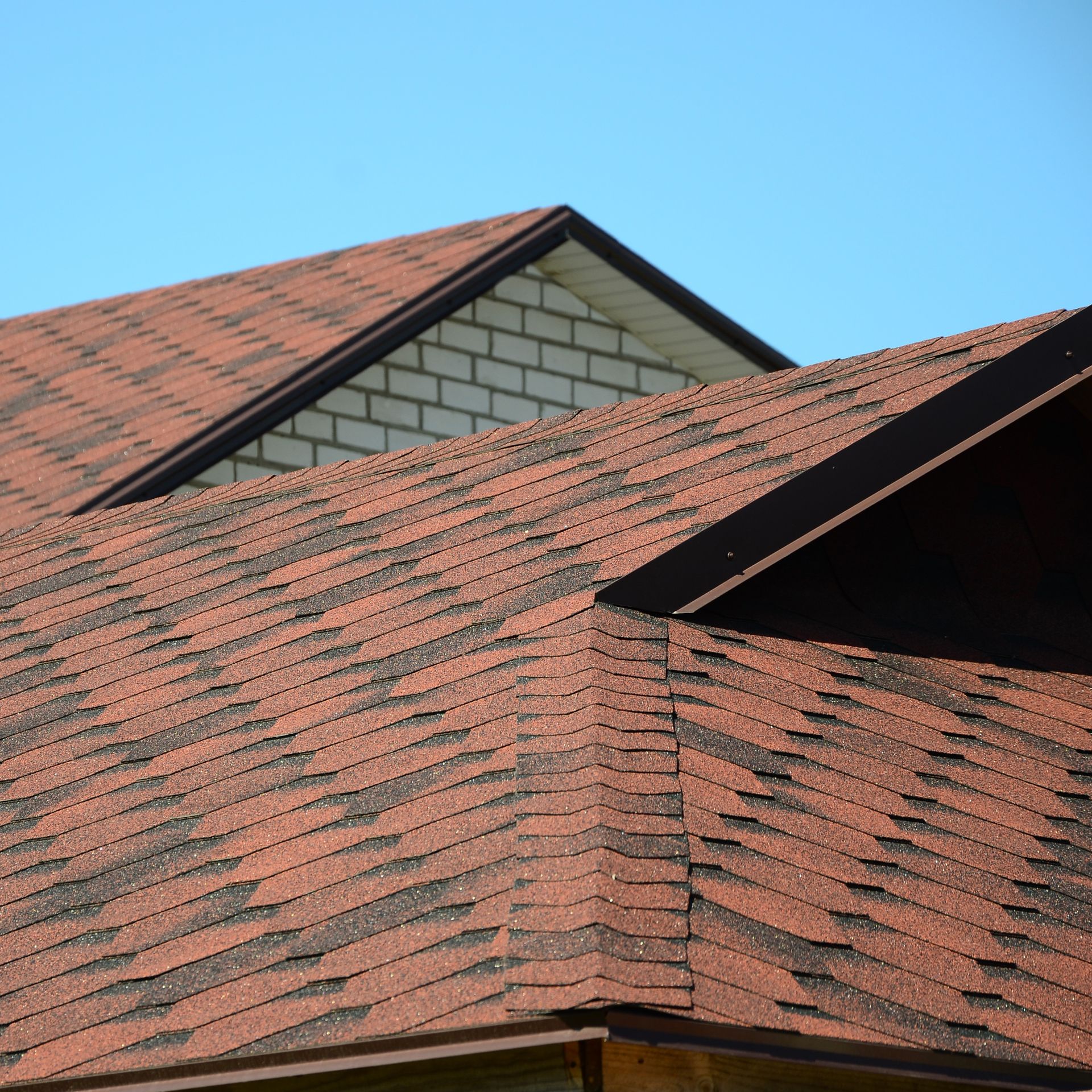 A close-up of a house roof with reddish-brown shingles and a white brick gable end against a clear blue sky.