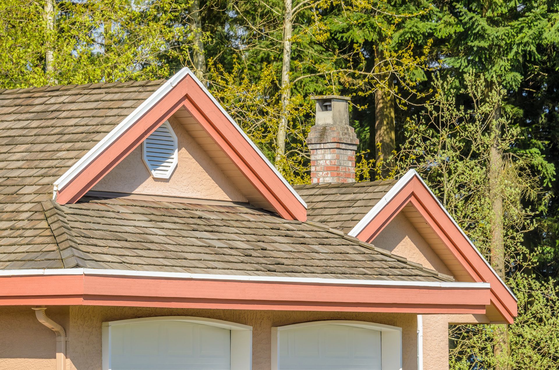 Tan roof with red trim, chimney, and peaked dormers, set against a blurred tree background.