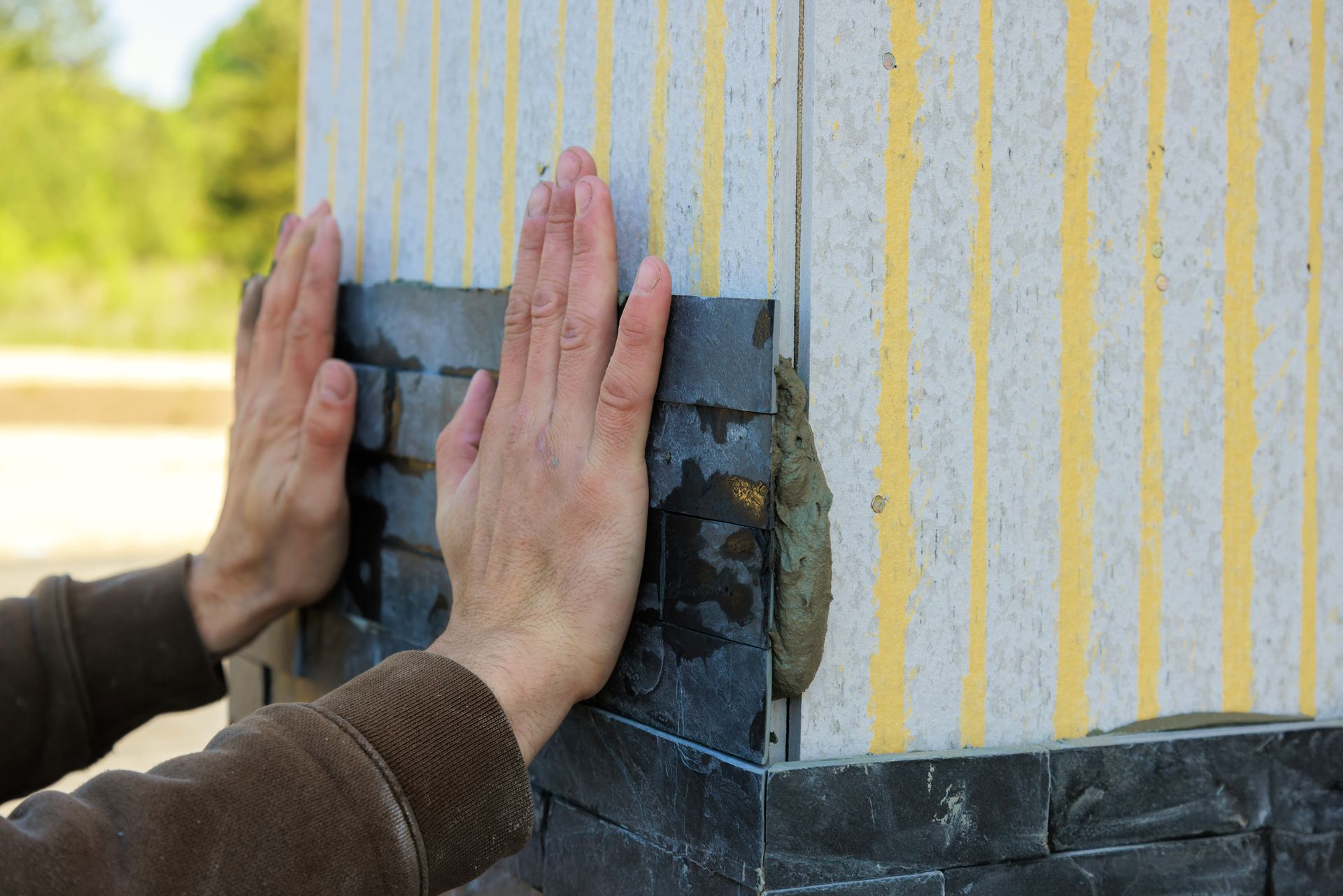 Hands press insulation board onto an exterior wall with yellow-striped siding and dark base trim.