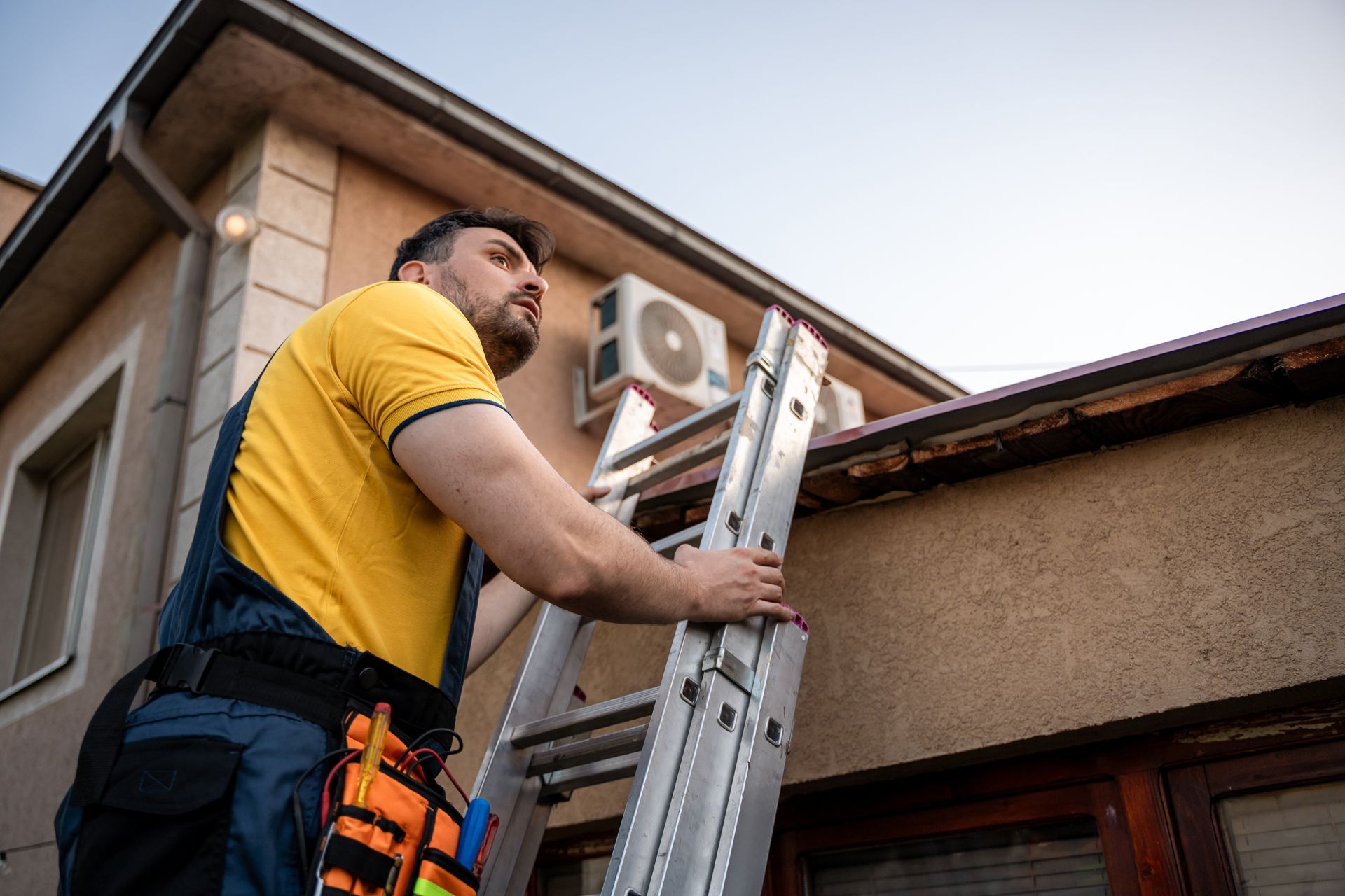 Man in yellow shirt climbs a ladder to inspect a building's roof and gutters.