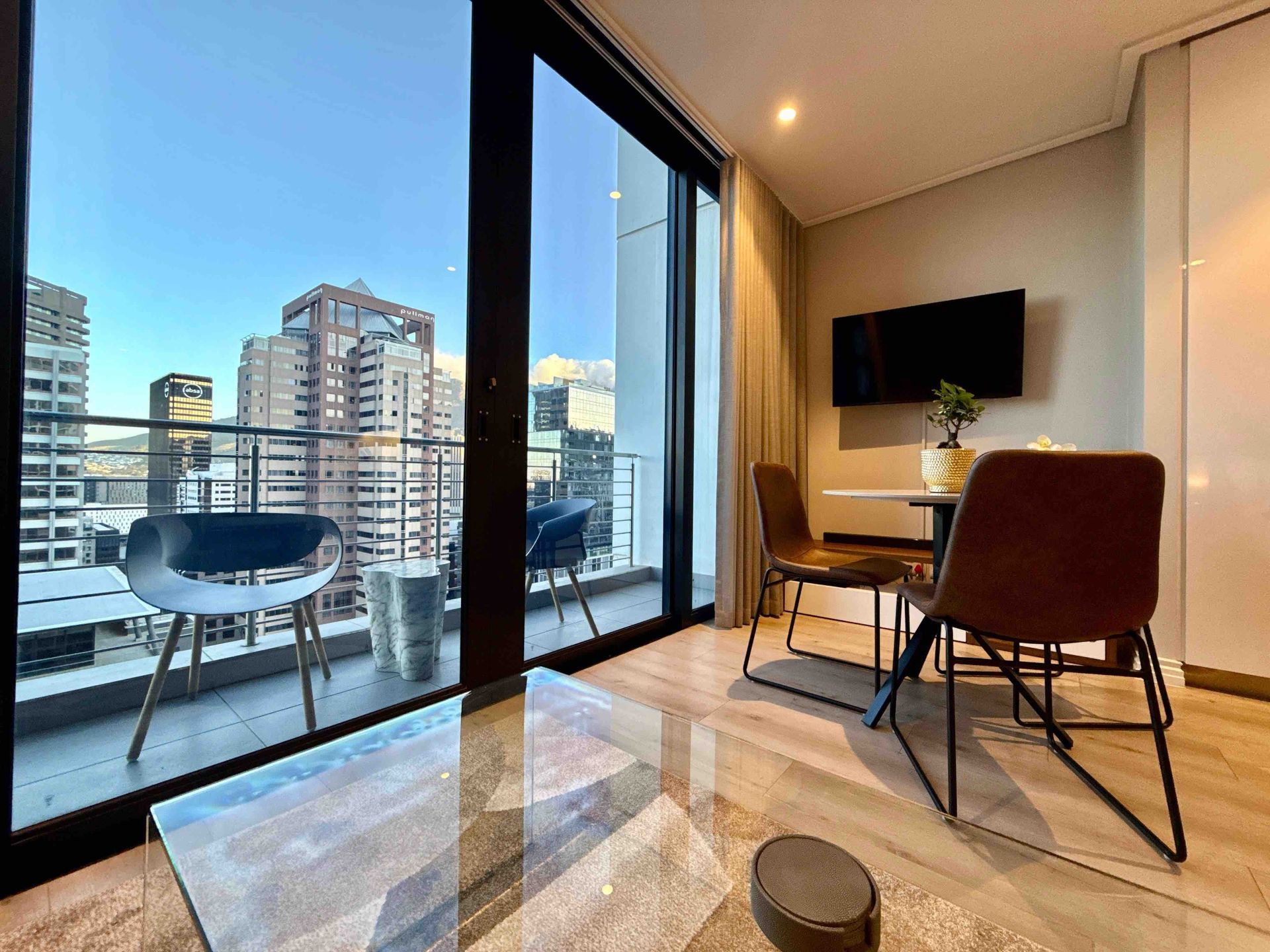 Balcony view from 16 on Bree Apartment 1906, showing outdoor seating, Cape Town city buildings, mountain views, and the open-plan living area with dining table and wall-mounted TV.