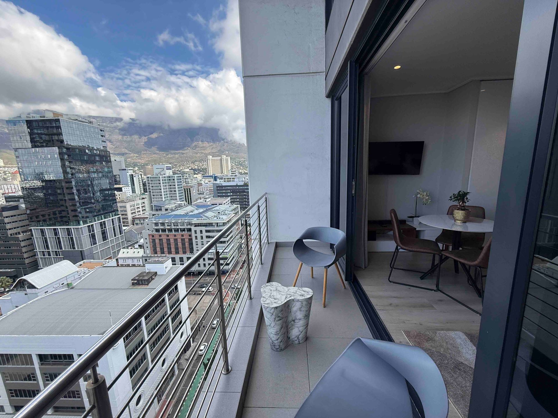 Balcony view from 16 on Bree Apartment 1906, showing outdoor seating, Cape Town city buildings, mountain views, and the open-plan living area with dining table and wall-mounted TV.