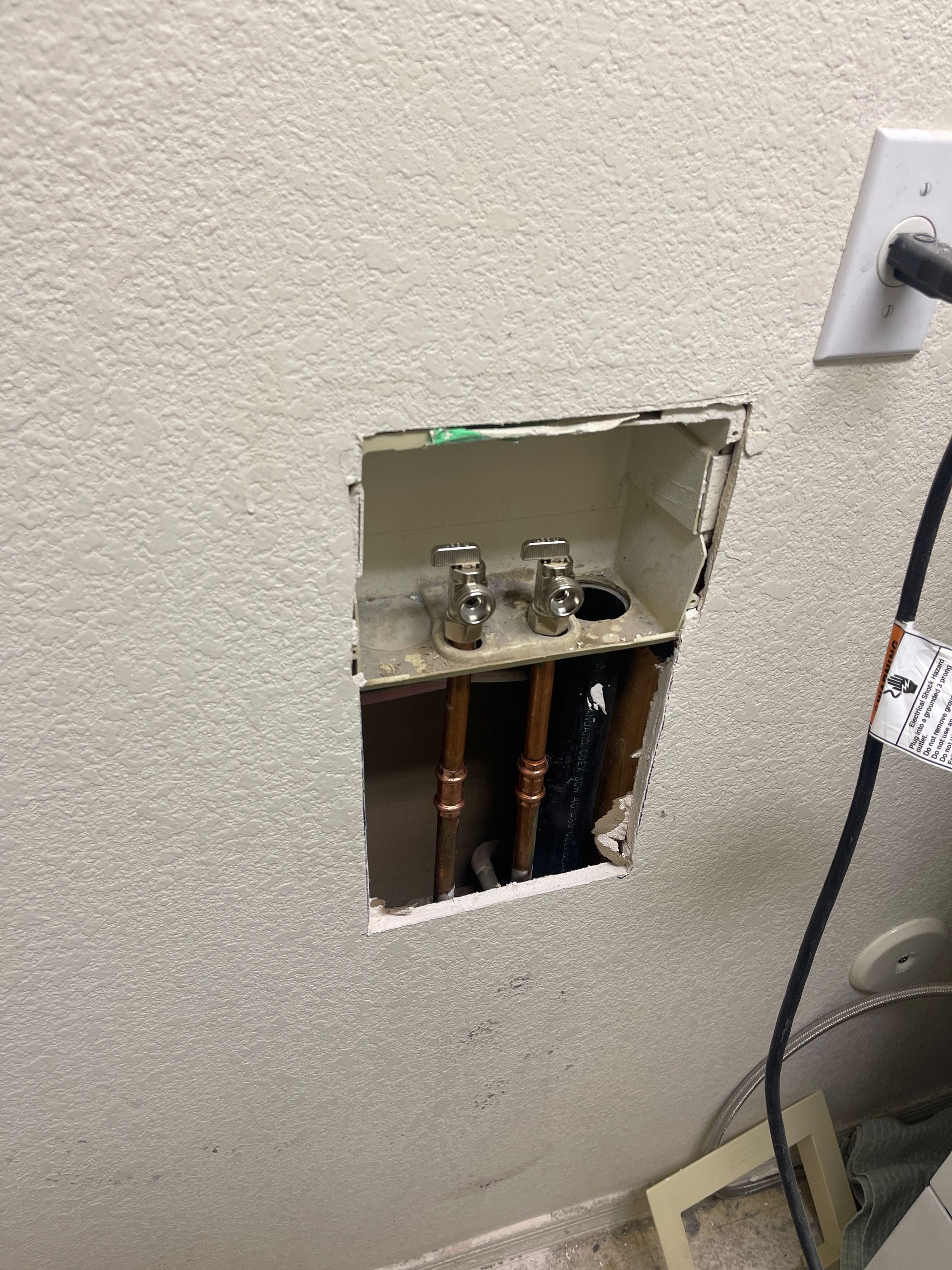 A hole in a textured wall reveals copper pipes and water valves for a washing machine, next to an electrical outlet.