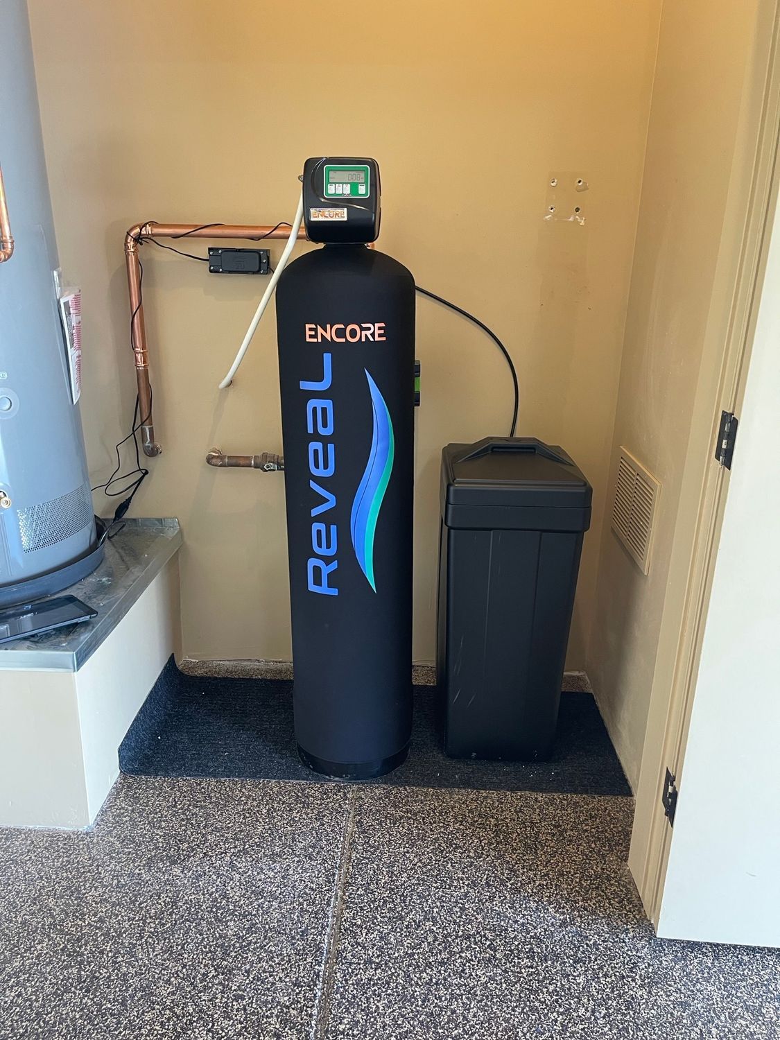 Water softener system with black tank and control unit, connected to plumbing in a utility room.