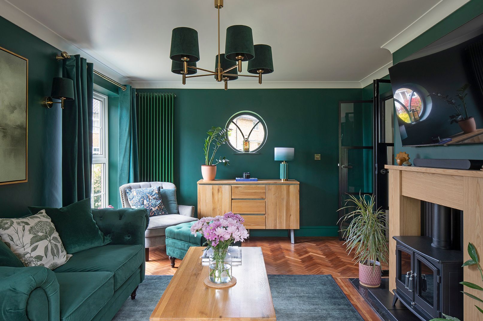 Hunter green living room with brass chandelier, bespoke oak shelving, and circular window in 1920s Welwyn Garden City home