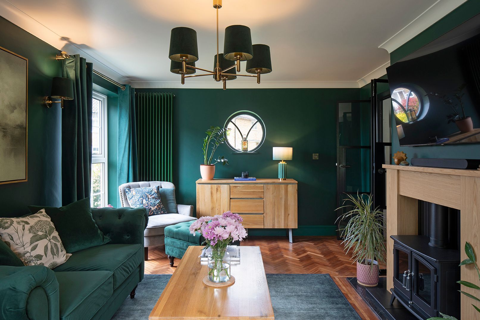 Hunter green living room with brass chandelier, bespoke oak shelving, and circular window in 1920s Welwyn Garden City home