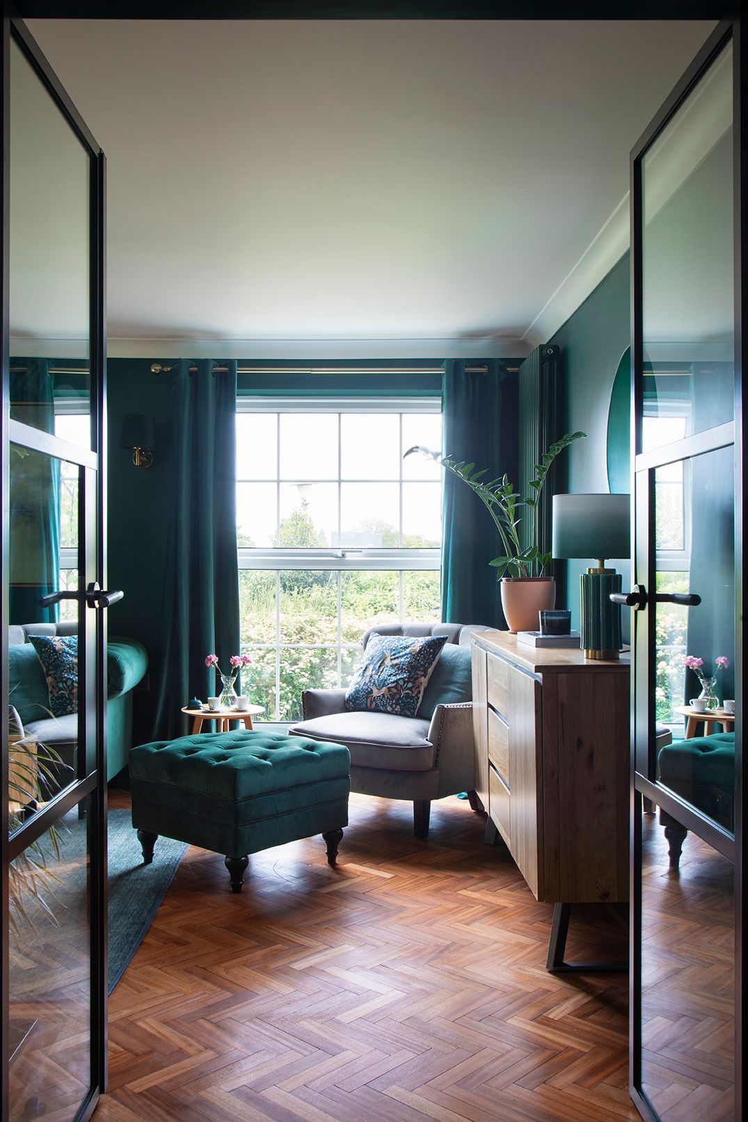 Black framed crittall doors opening to hunter green living room with herringbone oak flooring and bespoke joinery