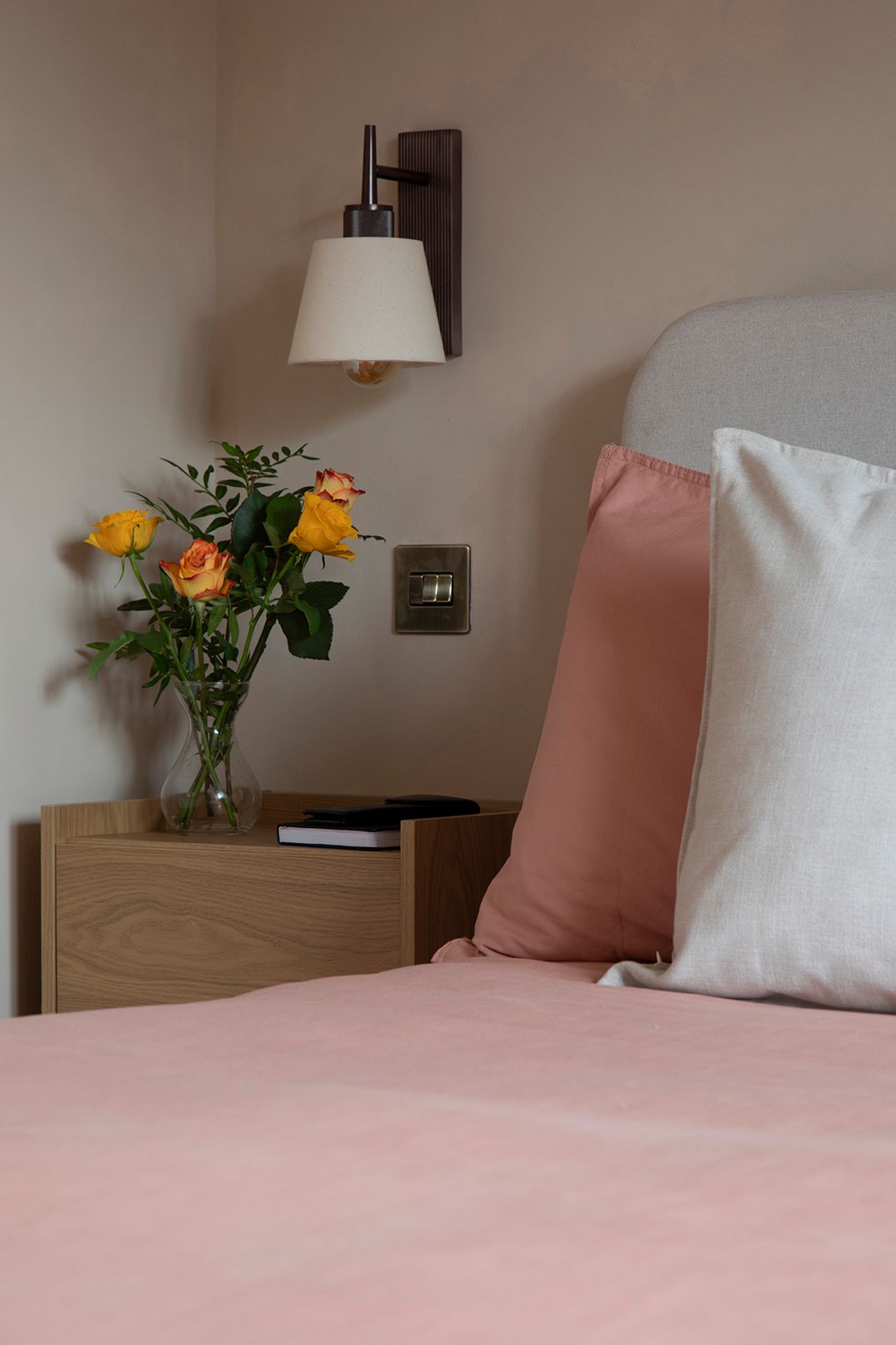 Close-up of oak nightstand with fresh orange roses and brass wall sconce — modern bedroom styling by Elisa Design in Watford.