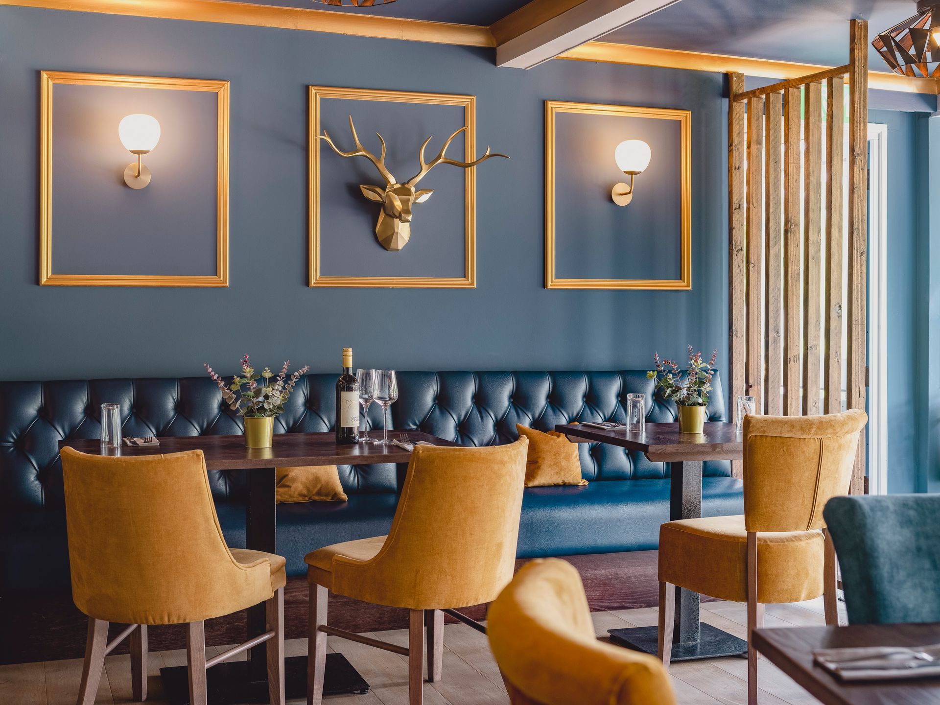 Golf clubhouse dining area with blue tufted banquette, mustard velvet chairs and gold stag head framed between wall lights.