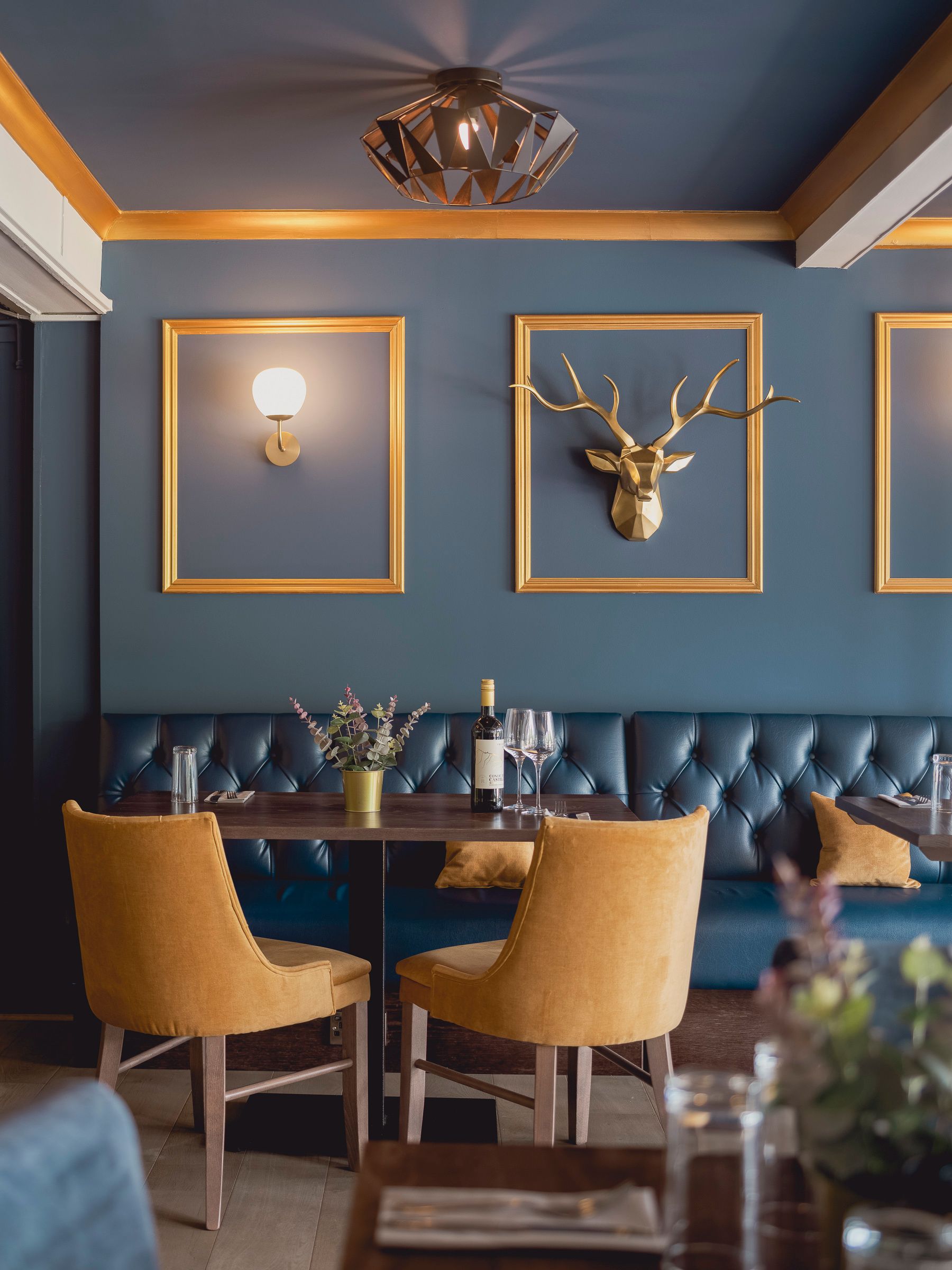 Golf clubhouse dining area with deep blue banquette, mustard velvet chairs and gold stag head on blue panelling.