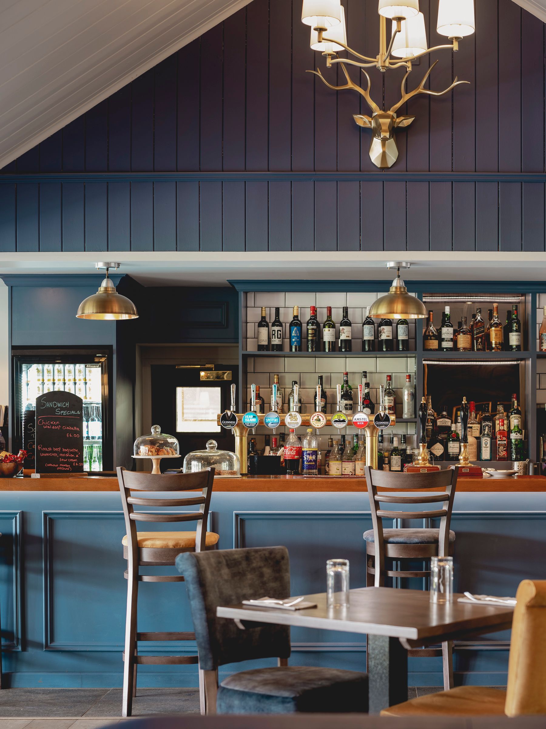Arkley golf clubhouse bar with blue panelled front, brass lighting, stocked back bar and gold stag chandelier above.