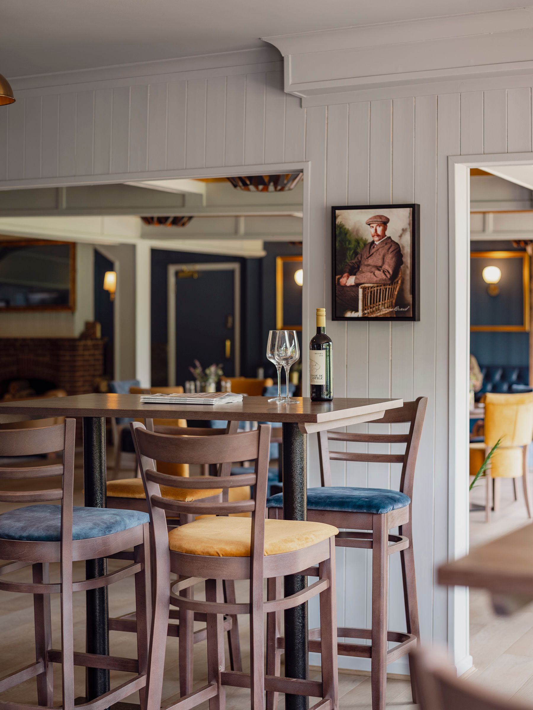 High dining table with blue and mustard bar stools, white panelling and vintage golfer artwork in Arkley golf clubhouse.