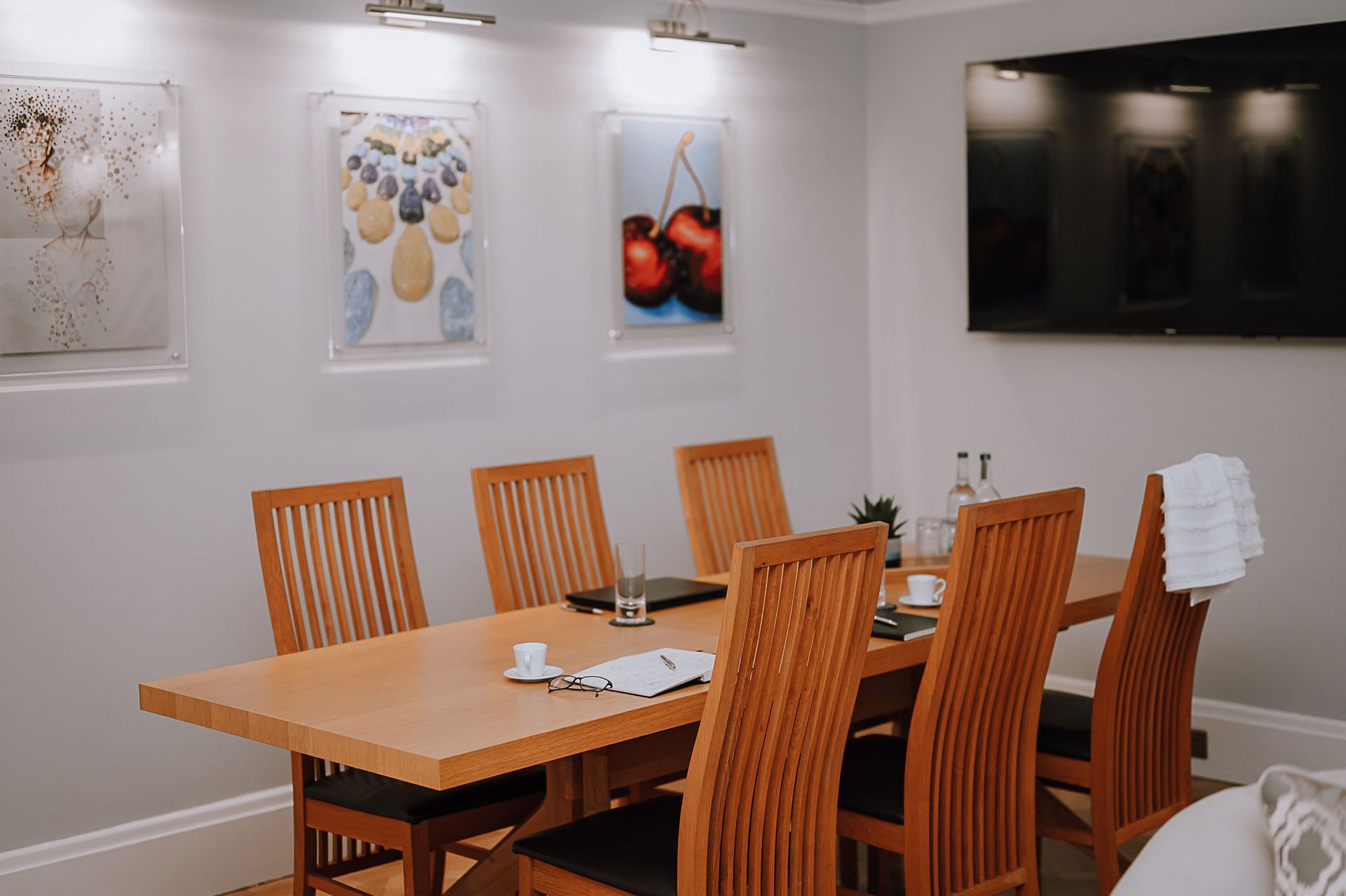 Meeting area with wooden table, high‑back chairs and pupil artwork lit by picture lights in Principal’s office.