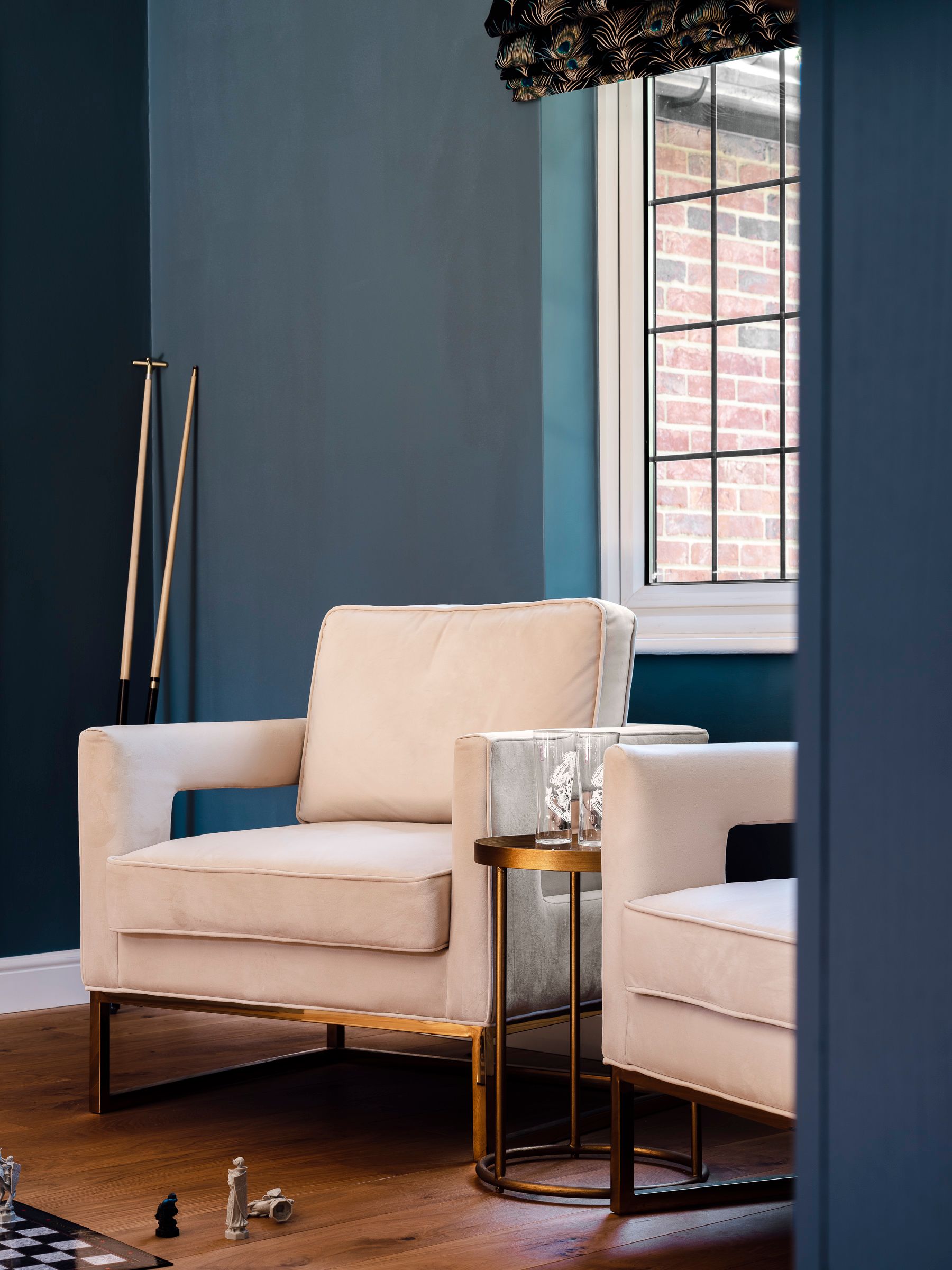 Games room seating area with cream armchairs, brass side table and blue walls with peacock‑print blind.