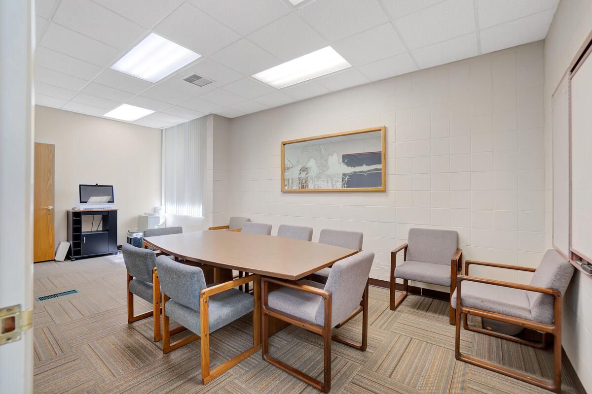 Meeting room with a rectangular table, chairs, artwork, and computer setup. Beige walls, carpet, and neutral furniture.