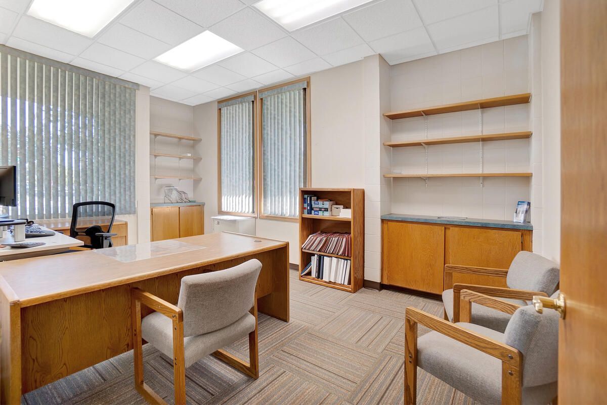 Office with a desk, two chairs, and built-in shelving with cabinets. Light brown furniture and beige carpet.