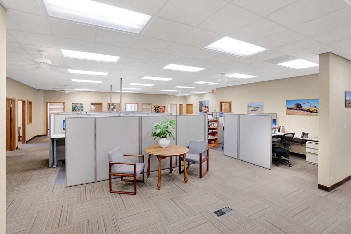 Office interior with cubicles, seating area, and overhead lighting. Beige carpet and gray cubicle walls.